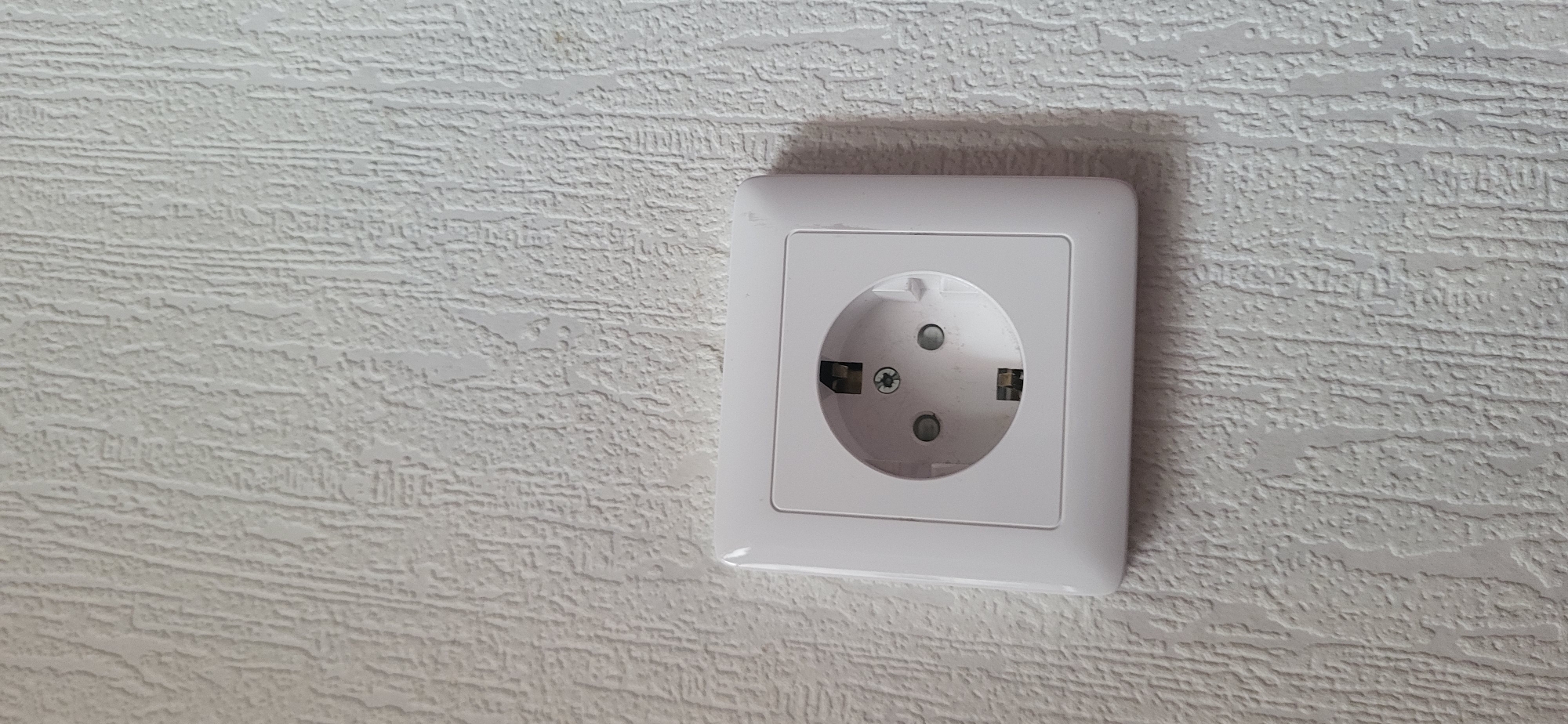 The image shows a white electrical outlet mounted on a textured white wall. The outlet is a European-style Schuko socket with two round holes and two grounding clips. The wall has a raised, linear texture, resembling wallpaper. The lighting is even, and there are no people or other objects visible in the frame. The context of Bulgakovo, Russia, is not visually evident from the image itself.