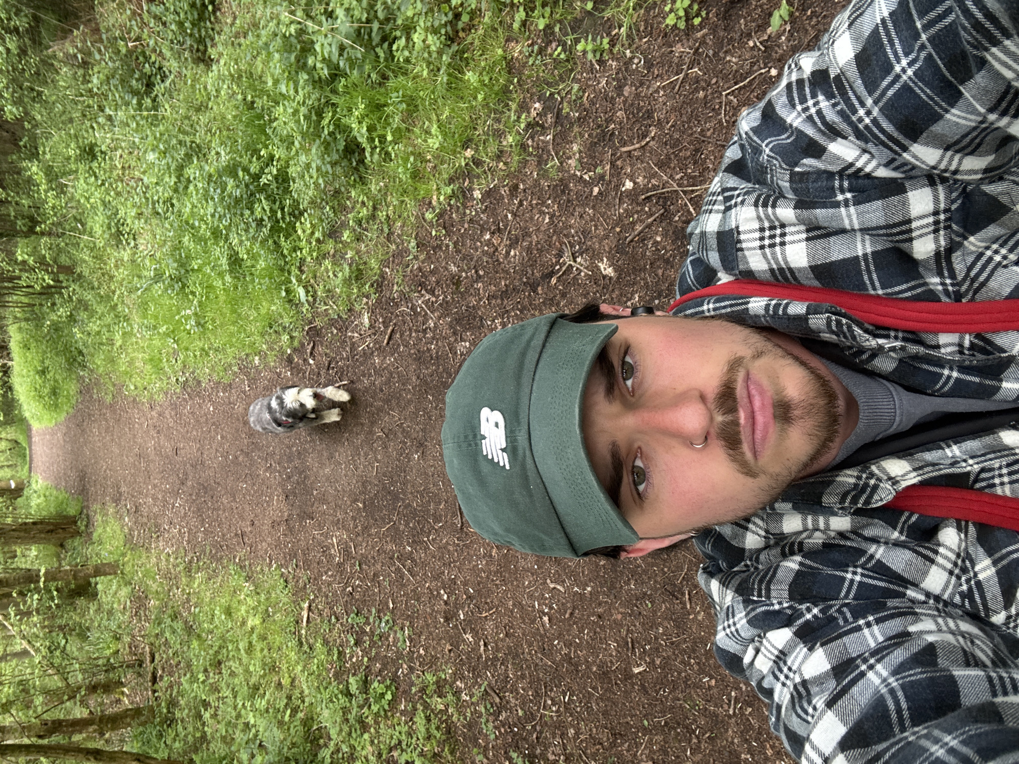 A man is posing for a selfie on a wooded path in an unknown city. He is wearing a grey and white plaid shirt, a grey undershirt, and a dark green New Balance baseball cap. He has brown eyes and a dark brown beard. Behind him, on the path, is a grey and white dog. The path is brown dirt and surrounded by green vegetation.