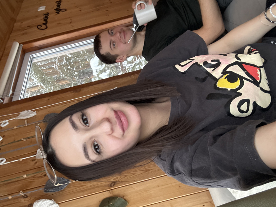 A selfie-style image depicts a young adult female in the foreground, smiling faintly towards the camera. She has long, dark hair, with light-framed spectacles perched on her head, resembling dark grey animal ears. She wears a dark grey t-shirt featuring a multi-colored cartoon graphic resembling the Pink Panther character. Her left arm is visible, adorned with a beaded bracelet on her wrist. In the mid-ground, to the right, a young adult male is seated, smiling at the camera while holding a white mug containing a metal straw or bombilla. He wears a dark t-shirt. They are indoors in a room with vertical light-brown wooden paneling on the walls. A large window behind the male reveals a snowy coniferous forest landscape. A light-colored roller blind is partially visible at the top of the window. Decorative elements resembling feather-like shapes and strung beads hang on the wall to the left of the window. To the right of the window, a dark cursive inscription reading 'Самый лучший' is affixed to the wooden wall. The scene is located in Sysert, Russia.