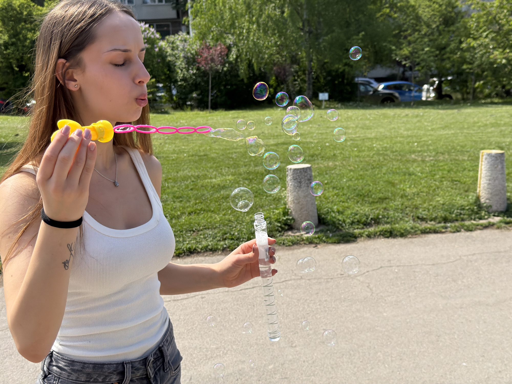 A young woman with long brown hair, wearing a white tank top and grey jeans, is blowing bubbles on a paved surface. She holds a yellow plastic bubble wand in her right hand and a clear plastic bubble solution container in her left. The background shows a grassy area with trees and buildings, suggesting a park. Based on the architecture and foliage, the location is likely in the city of Sofia, Bulgaria. Several bubbles float in the air, and two stone posts are visible in the background. The woman's eyes are closed as she blows through the bubble wand.