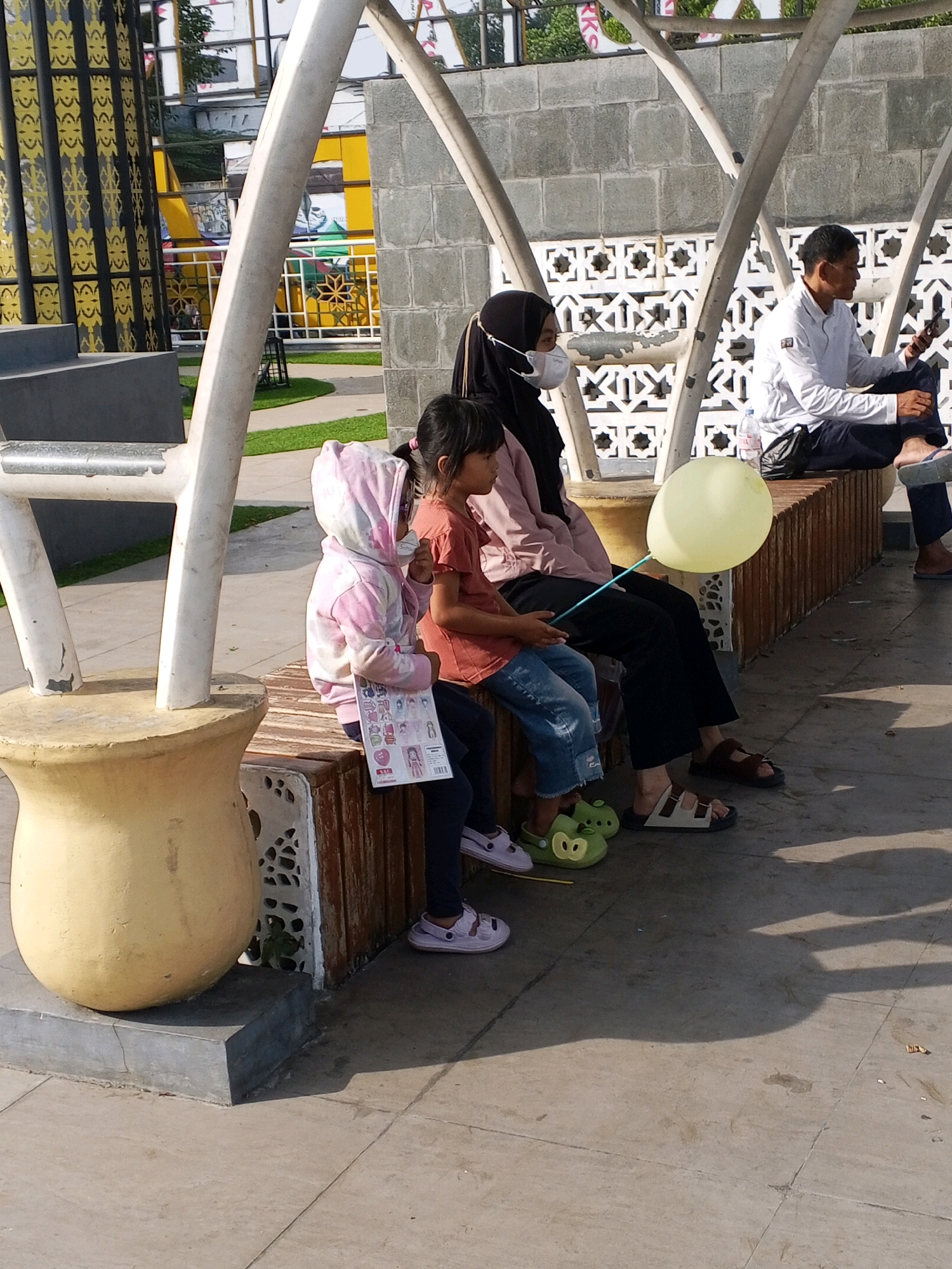 In Serang, Indonesia, a group of people are seated on a wooden bench outdoors, under a white canopy structure. A woman in a black hijab and a face mask is seated with a child. To her right, another child is seated. In front of them, a yellow balloon is attached to a stick. A man in a white shirt is seated to the right of the woman, looking at his phone. The ground is paved with light-colored tiles. To the left of the bench, a large, yellow, vase-shaped object is visible. The background features a brick wall with decorative patterns and a hint of greenery.