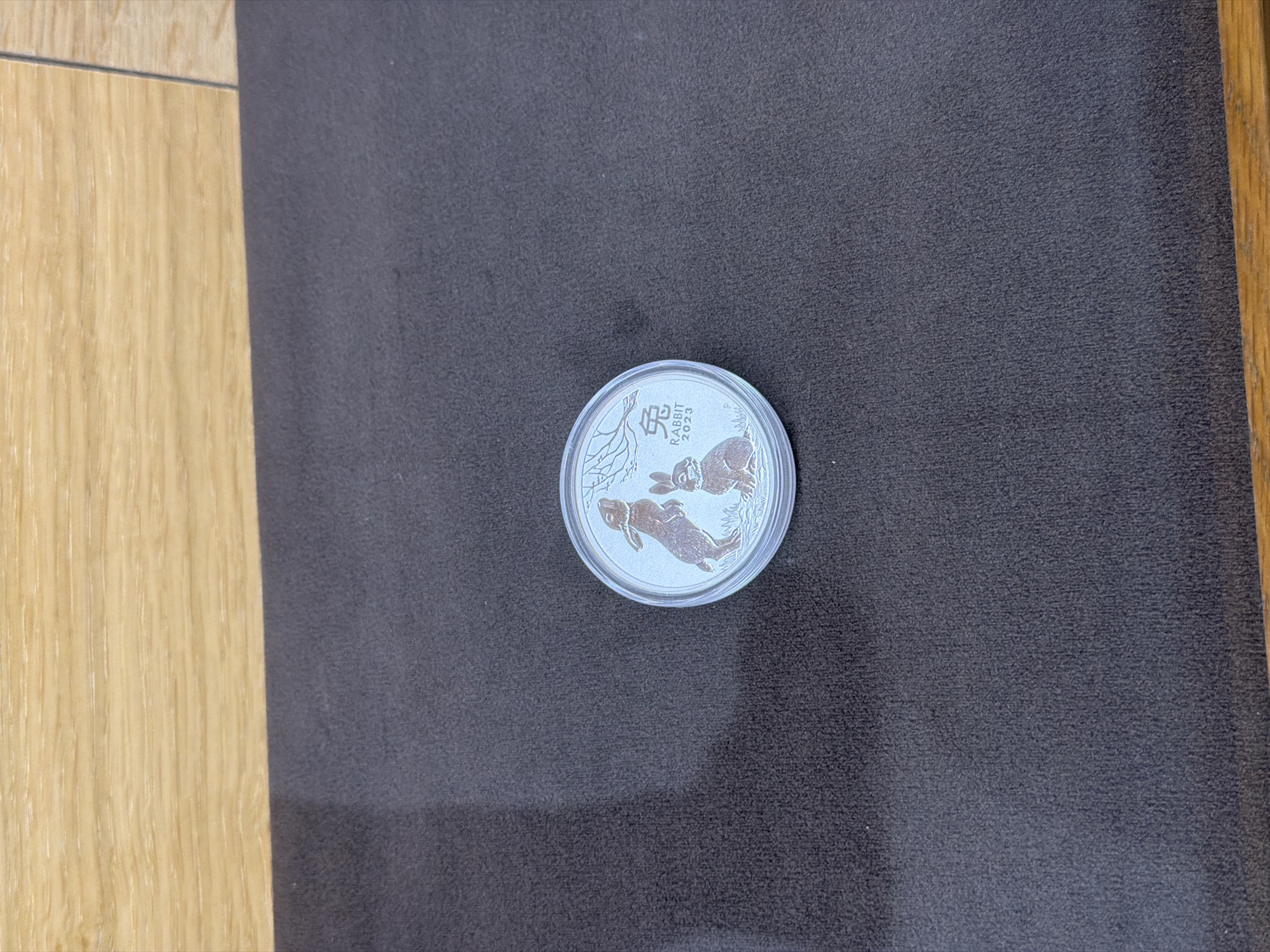 A silver coin encased in a clear plastic protector rests on a dark grey, textured surface. The coin features two rabbits beneath branches on one side, with the Chinese character for "Rabbit" and the year "2023" inscribed above. To the left, a light-colored wooden surface extends vertically. The scene is indoors. There is no indication of the location being Burgas, Bulgaria.