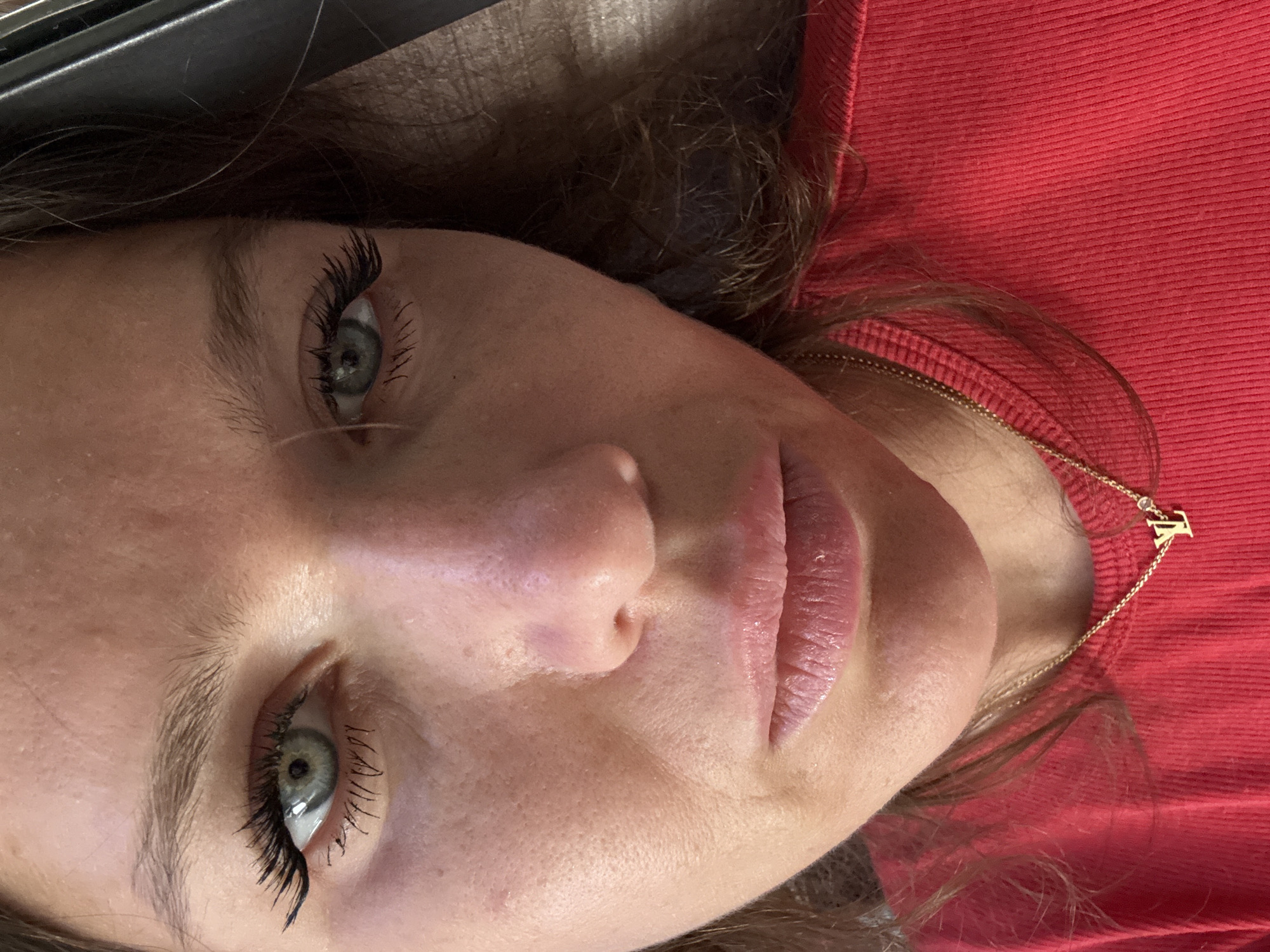 A close-up, angled portrait of a woman's face. She has light skin, blue-green eyes with dark mascara, and pink lips. She is wearing a red ribbed shirt and a gold necklace with a "T" pendant. The background is out of focus. The location is not discernible from the image.