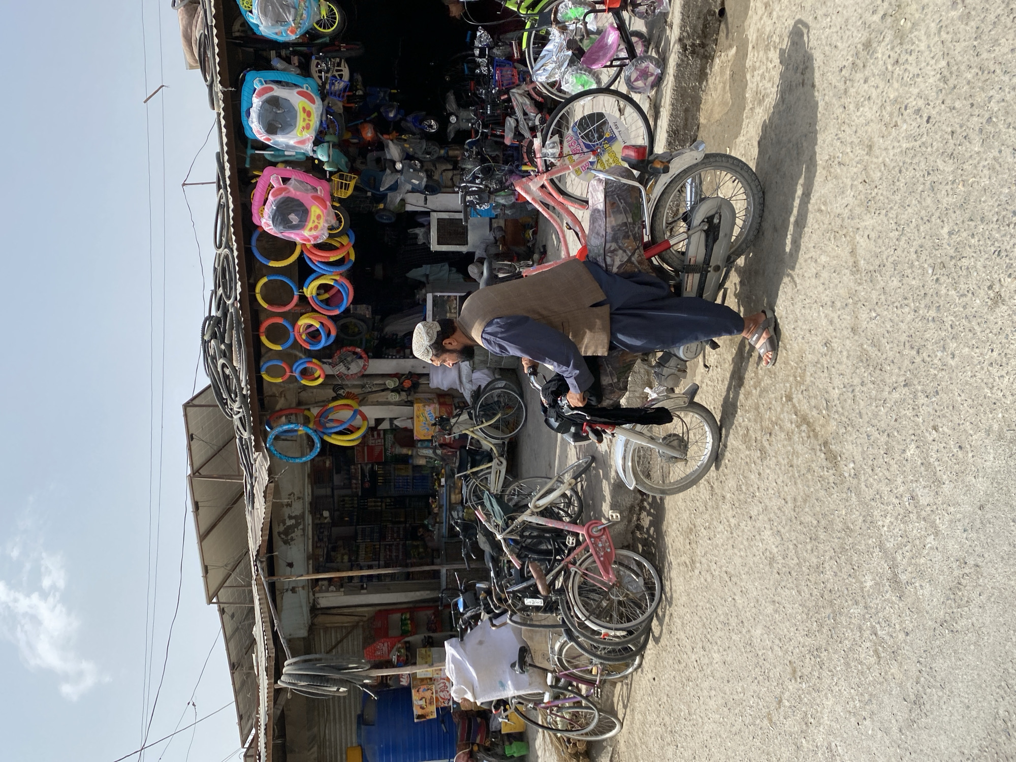 The image depicts a street scene in Kandahar, Afghanistan, featuring a man on a motorcycle in front of a shop that sells bicycles and related items. The shop's facade is lined with various colorful bicycle wheels, tires, and other accessories hanging from the roof. Behind the man, a collection of bicycles, including children's bikes and adult models, are parked. The man is wearing traditional Afghan attire, including a vest and a skullcap. The ground is a mixture of concrete and gravel. The overall atmosphere suggests a commercial area within the city.