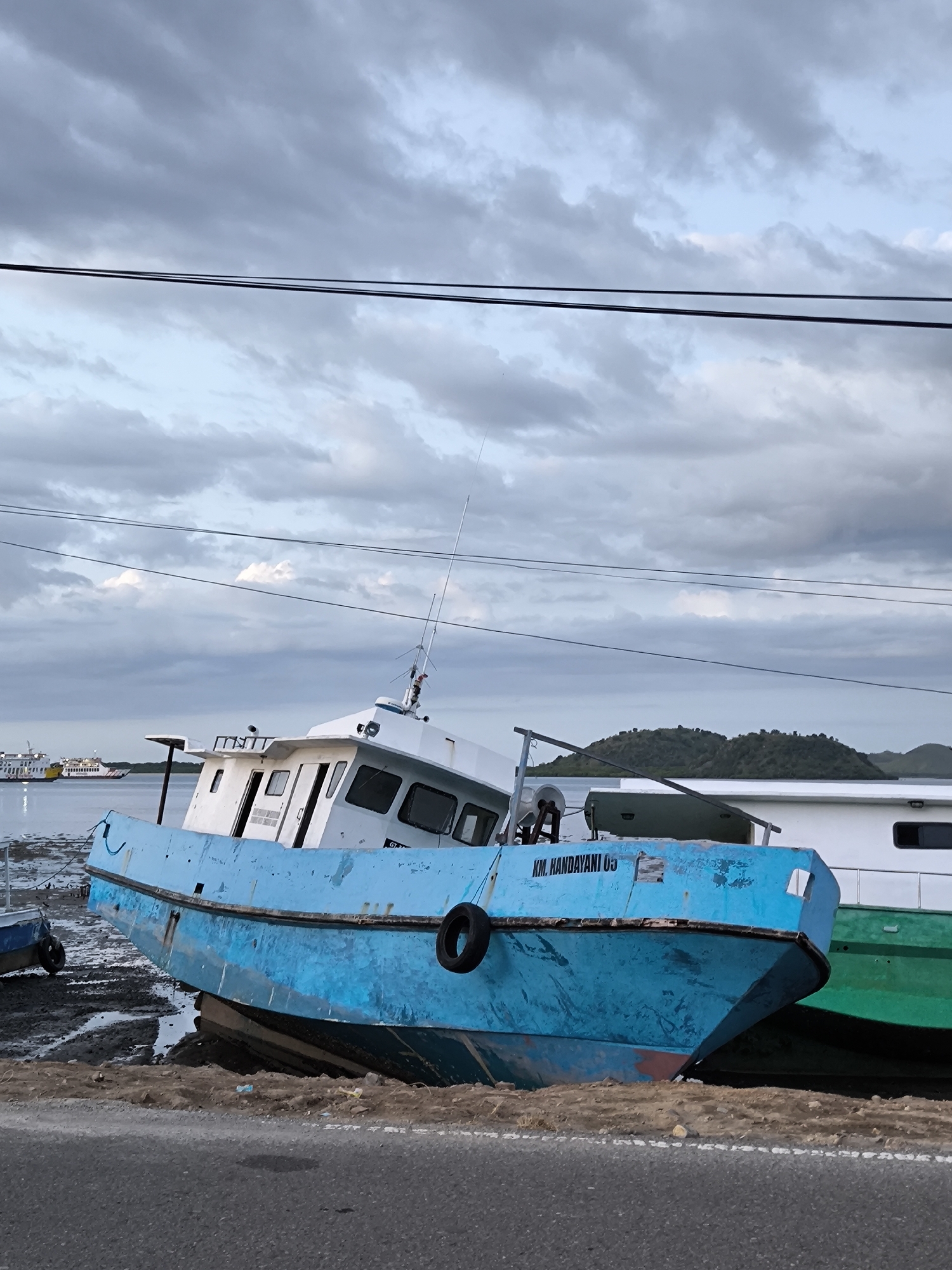The image shows a blue boat named "KM. HANDAYANI 03" beached on the shore in Upt Tambak Sari, Indonesia. The boat leans to the left. A black tire is attached to its side. Mud and sand surround the boat's hull. To the right, parts of another boat can be seen, with white and green sections visible. In the background, there is water, a distant landmass, and a cloudy sky. A road runs along the bottom of the frame. Power lines are strung across the upper portion of the image.