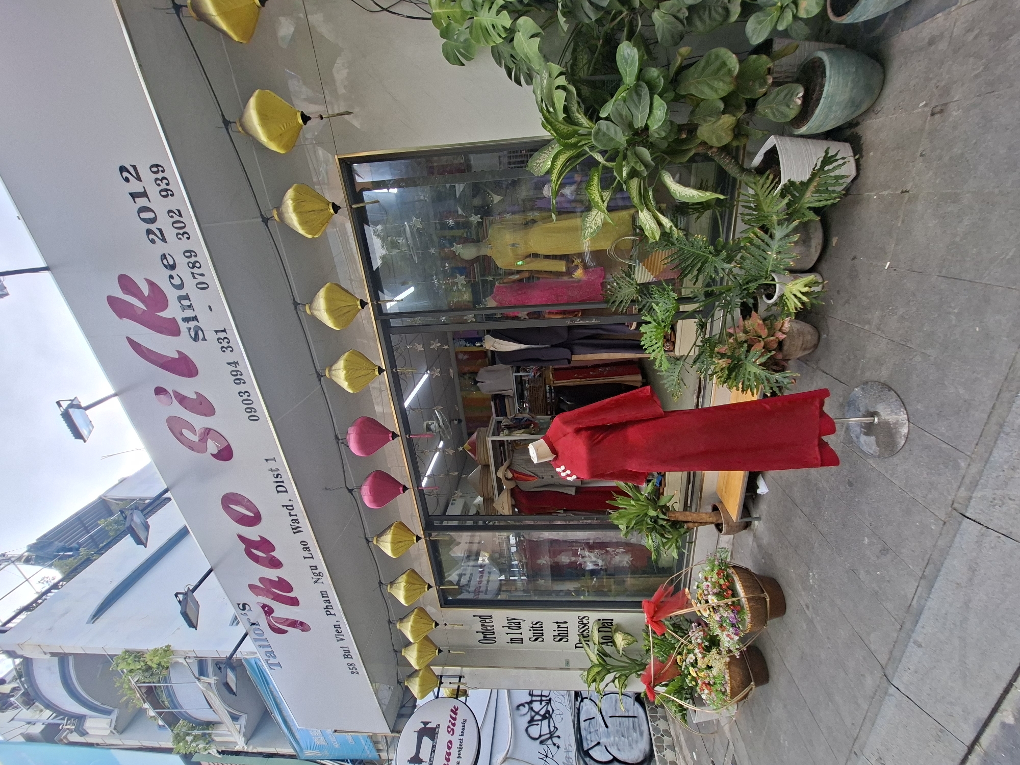 The image depicts the storefront of a tailor shop named "Thao Silk" located at 258 Bui Vien, Pham Ngu Lao Ward, District 1, city of Thủ Đức, Vietnam. The shop's name is prominently displayed on a white awning with "Thao Silk" written in a large, stylized pink font, accompanied by the text "since 2012" and phone numbers. Red and yellow lanterns hang in front of the shop.
The shop's interior is visible through large glass windows, showcasing mannequins dressed in traditional Vietnamese attire and suits. Plants are arranged in pots along the sidewalk in front of the store, adding greenery to the urban environment. Two decorative baskets filled with flowers are placed near the entrance. The sidewalk is paved with grey tiles. In the background, a multi-story building with balconies and some graffiti is visible. The sky is bright, suggesting it is daytime. There are no people clearly visible in the image.
