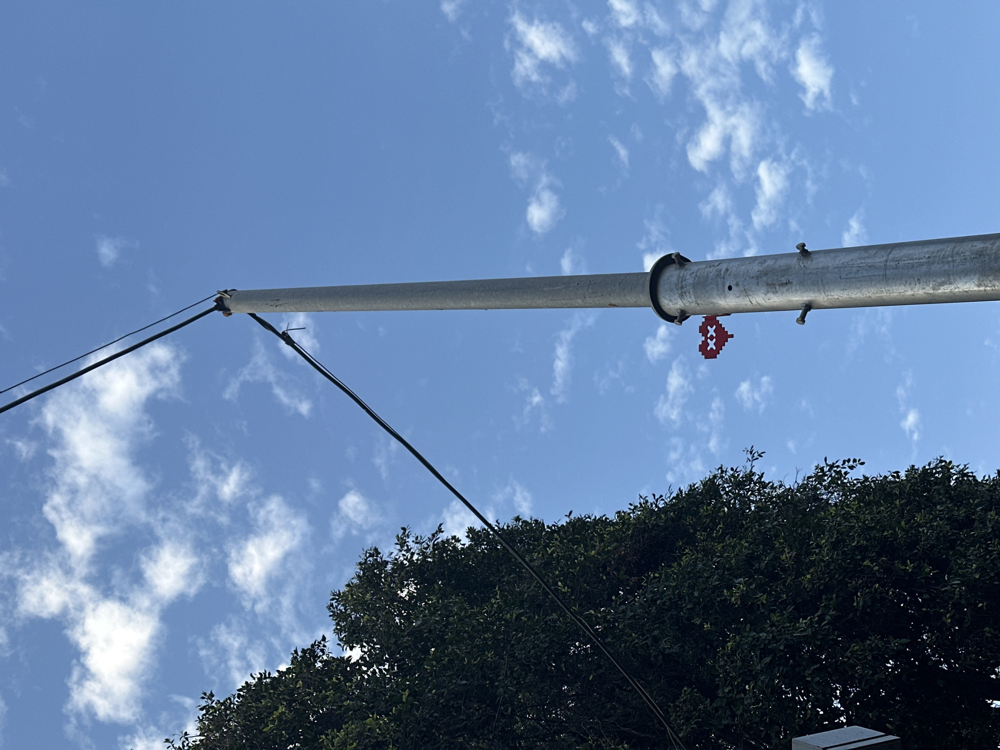 The image presents a low-angle shot of a utility pole extending towards a blue sky with scattered clouds. The pole is grey, metallic, and features several visible bolts and attachments. A red pixelated heart-shaped object is affixed to the pole. Two black cables are connected to the pole, stretching diagonally across the frame. The lower portion of the image includes the tops of a dense green tree and what appears to be a street lamp. The location, Tel-Aviv, Israel, is not discernible from the image content.