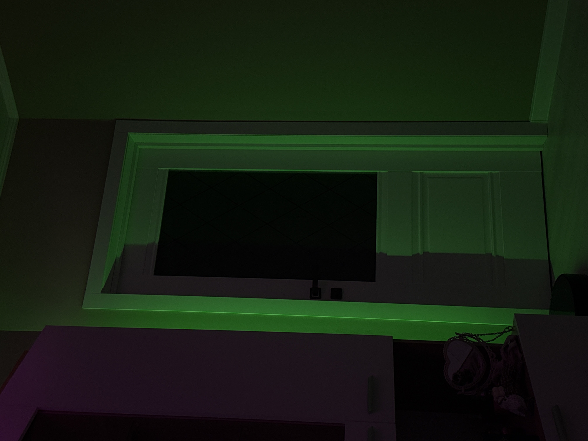 A low-angle, indoor shot presents a dimly lit room. A flat-screen television, unlit, is mounted within a white frame, flanked by what appears to be a set of cabinet doors or wall paneling. The lower portion of the image reveals a pinkish-purple cabinet with what look like metallic handles. The scene is bathed in a green glow, likely from LED strip lighting positioned around the frame of the television. To the right, a hint of miscellaneous objects and a partial view of another white cabinet can be seen.