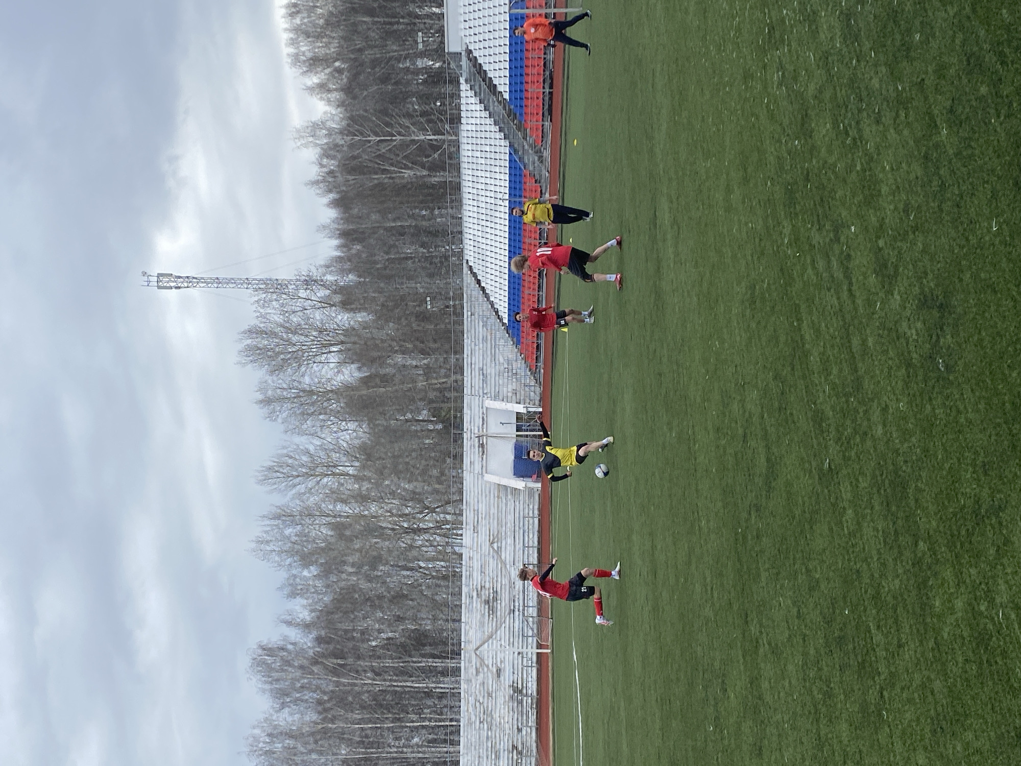 The image shows a soccer practice or game taking place in Chaikovsky, Russia, likely at the Central Stadium (Центральный стадион). The foreground features a green soccer field with several players. Some players are wearing red and black uniforms, while another player is wearing a yellow and black uniform. One player in yellow is kicking the soccer ball. Other players are running on the field. Behind the field is stadium seating with sections painted in the colors of the Russian flag: red, white, and blue. Beyond the stadium seating, a row of trees without leaves is visible against a gray, overcast sky. A tall metal communications tower is also visible rising behind the trees.