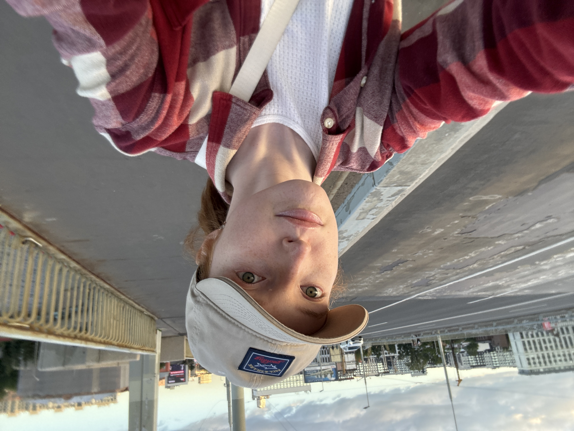 A young woman with fair skin, freckles, and red hair is taking a selfie on a bridge in a city. She is wearing a tan baseball cap with a logo, a white waffle-knit shirt, and a red and white plaid flannel shirt. A white strap is visible across her chest. She is looking at the camera with a neutral expression. The background shows a road with cars and buildings on either side, and a railing along the side of the bridge. The sky is cloudy.