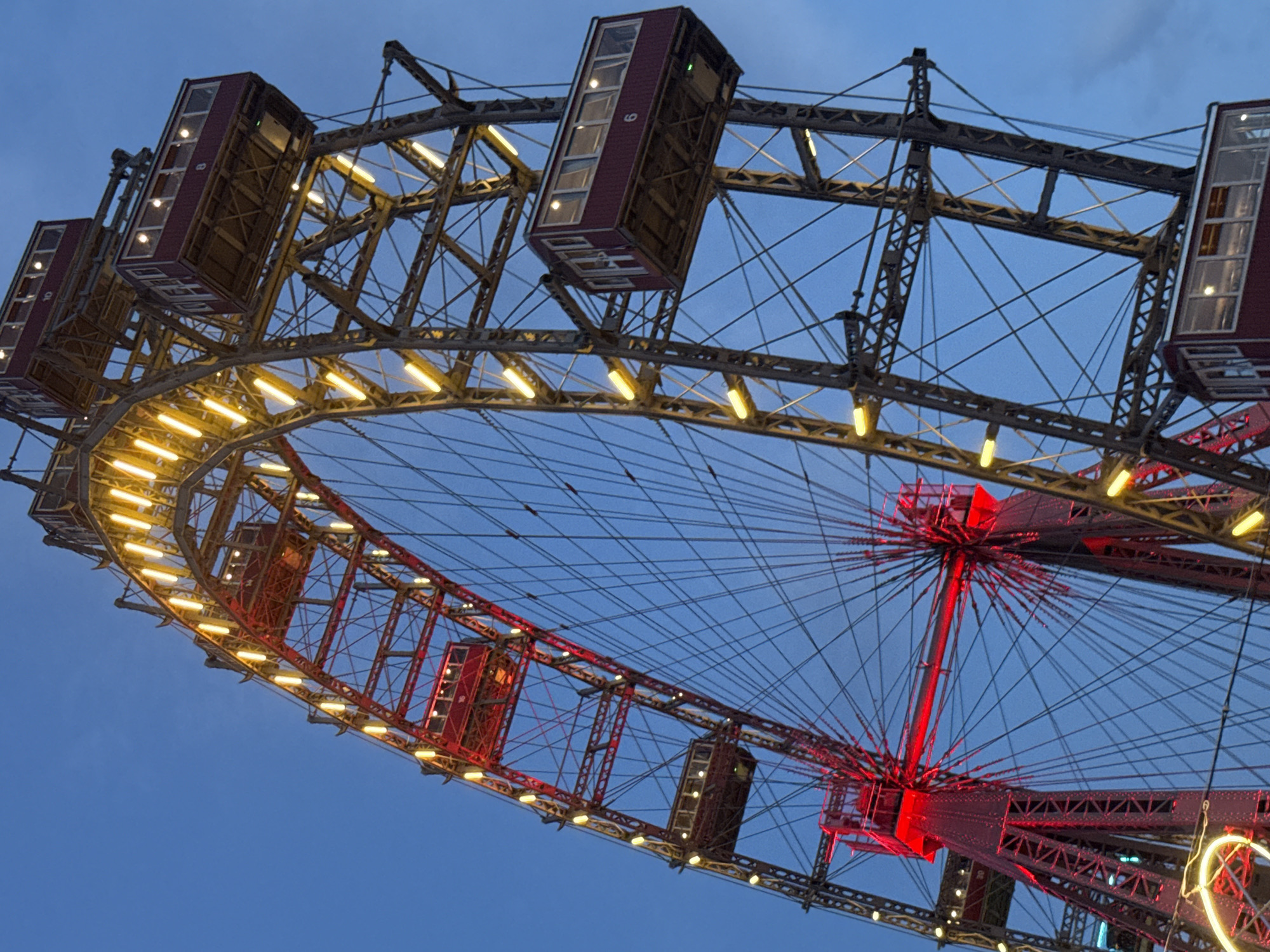 This is a low-angle, close-up, vertical, full-shot photograph of the Wiener Riesenrad, a historic Ferris wheel located in the Prater amusement park in Vienna, Austria. The sky is a deep, twilight blue. The Ferris wheel's structure is made of dark metal, with crisscrossing beams and cables. It is illuminated by a combination of red lights on the lower support structure and yellow lights along the outer rim of the wheel. Several of the Ferris wheel's enclosed, maroon-colored gondolas are visible, each with windows showing interior lights. One gondola prominently displays the number "6" on its side. The overall scene depicts the iconic landmark at dusk, bathed in artificial light against the darkening sky.