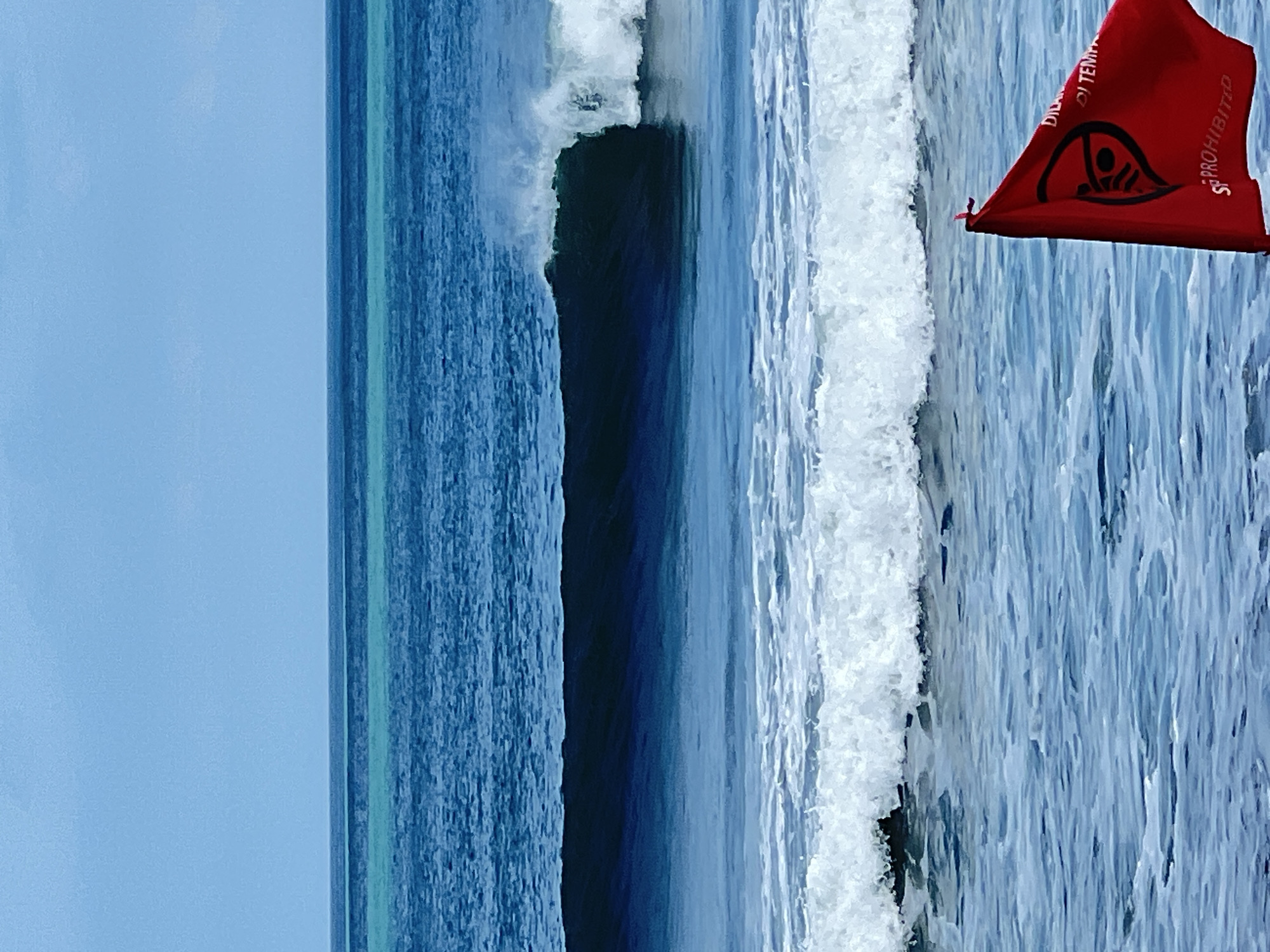 A bright red flag with black symbols and text is planted in the shallow, rippling ocean water in the foreground of the image. The flag displays a circle with a diagonal line through it, indicating prohibition, and appears to have text in Indonesian, with "DILARANG" and "DIPROHIBITED" visible. Behind the flag, the ocean extends to the horizon, with multiple layers of waves. A large, dark wave is breaking in the middle ground, casting a shadow. Further out, the ocean transitions to lighter blues and turquoise hues, meeting a pale blue sky. The scene depicts a beach environment, likely Kuta, Indonesia, with a warning flag indicating potentially dangerous conditions or restricted activity in the water.