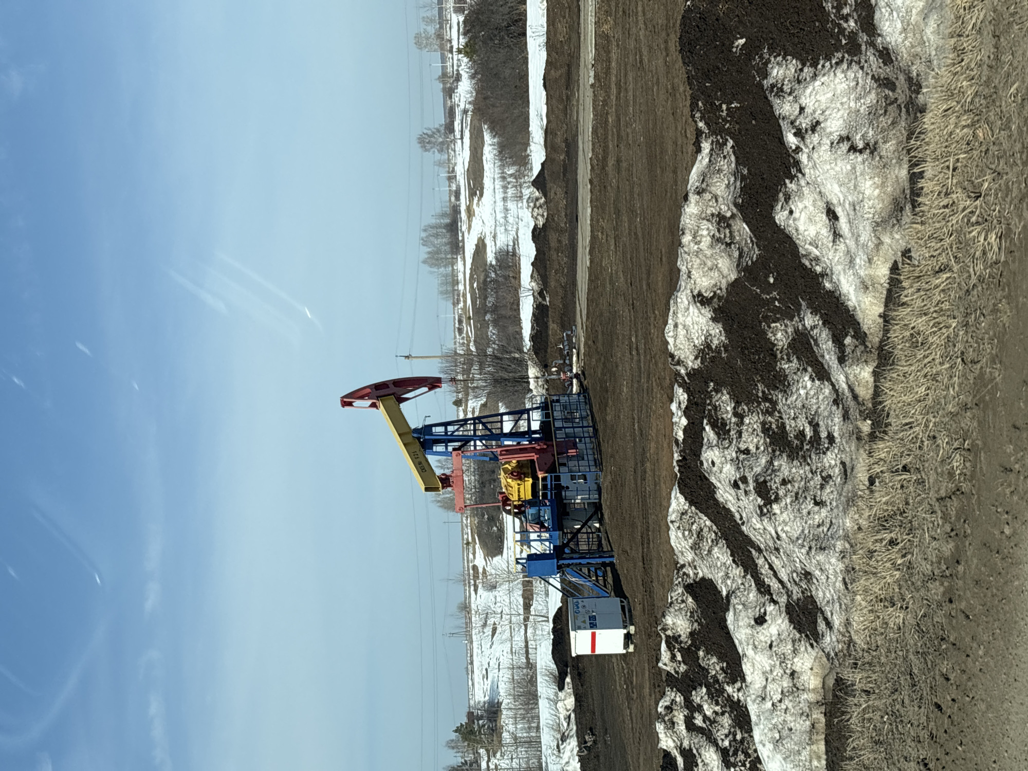 The image shows an oil pump jack in a rural setting, likely located near Ustinovo, Russia. The pump jack is painted with red, yellow, blue, and black details, and is situated in a snowy field interspersed with bare trees and vegetation. The ground is a mix of brown earth and patches of melting snow. A utility box with red markings sits adjacent to the pump. The sky above is clear and blue. The scene appears to be taken during the daytime.