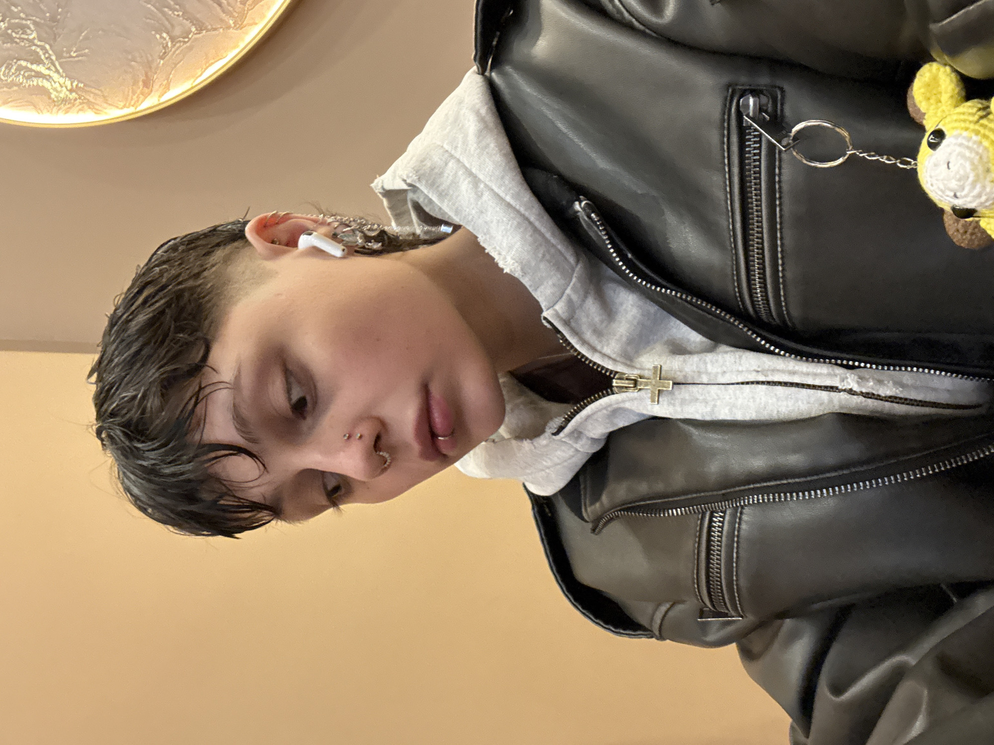 A close-up, slightly rotated shot features a person with short, dark, wet-look hair and a modern hairstyle. They have piercings on their nose and ear, and an earbud is visible in their ear. The person is wearing a grey hoodie with a cross design on the zipper pull, layered underneath a black leather jacket. A small, crocheted yellow animal charm is attached to the jacket's zipper. The background is a plain, light tan color, with a circular decorative piece partially visible in the upper left corner.