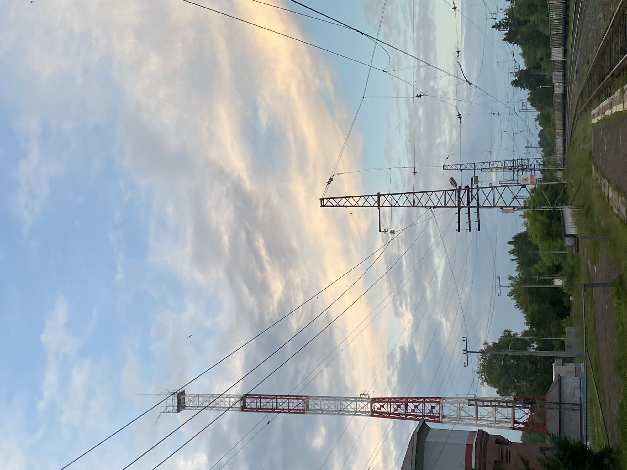 The image depicts a railway scene at what is identified as Borodulino, Russia. Dominating the foreground are railway tracks curving towards the right of the frame. Along the tracks, a platform is visible. To the left, a large water tower stands, adjacent to a tall, red and white lattice tower, likely a telecommunications mast. Several other similar lattice towers are visible in the distance, along with overhead power lines that crisscross the sky, indicating an electrified railway. A dense line of trees borders the railway on the right. The sky is filled with a mix of blue and white clouds, with hints of sunset colors. Several birds are visible in flight. The overall impression is of an industrial or semi-industrial area on the outskirts of a town.