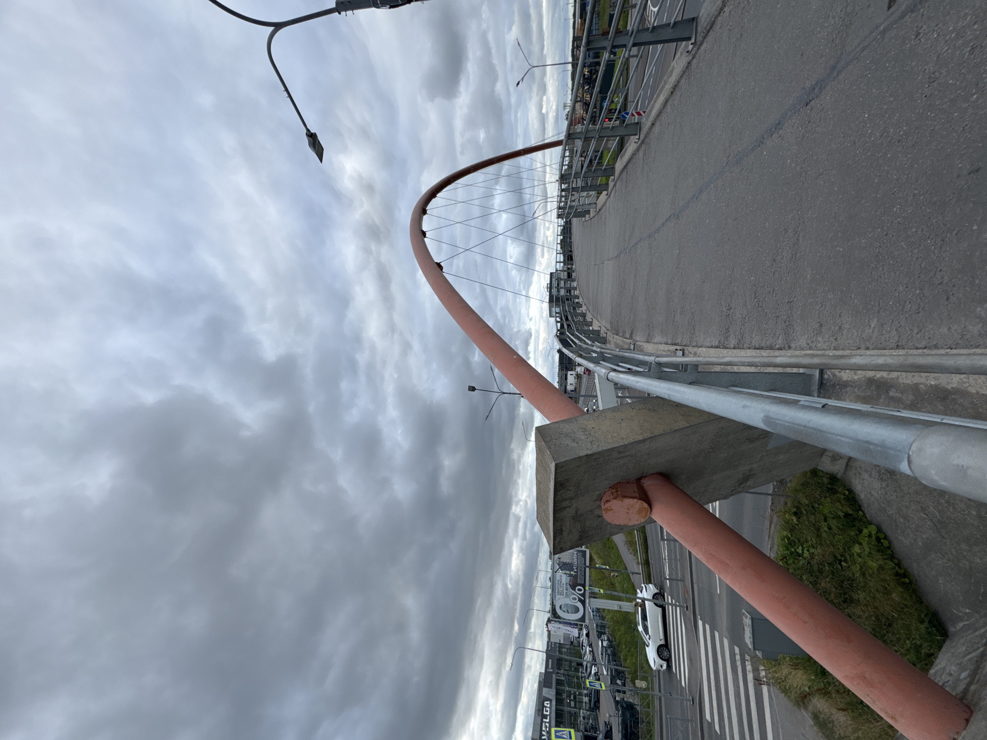 The image shows a pedestrian bridge in Saint Petersburg, Russia, under a cloudy sky. The bridge has a distinctive orange arch and is supported by cables. In the foreground, a large, weathered concrete block with a red bolt protrudes from the bridge structure. A pedestrian walkway is visible, curving to the right. Below the bridge, a street with a white car and a pedestrian crossing can be seen. In the background, buildings and streetlights are visible. The overall scene suggests an urban environment on a gloomy day.