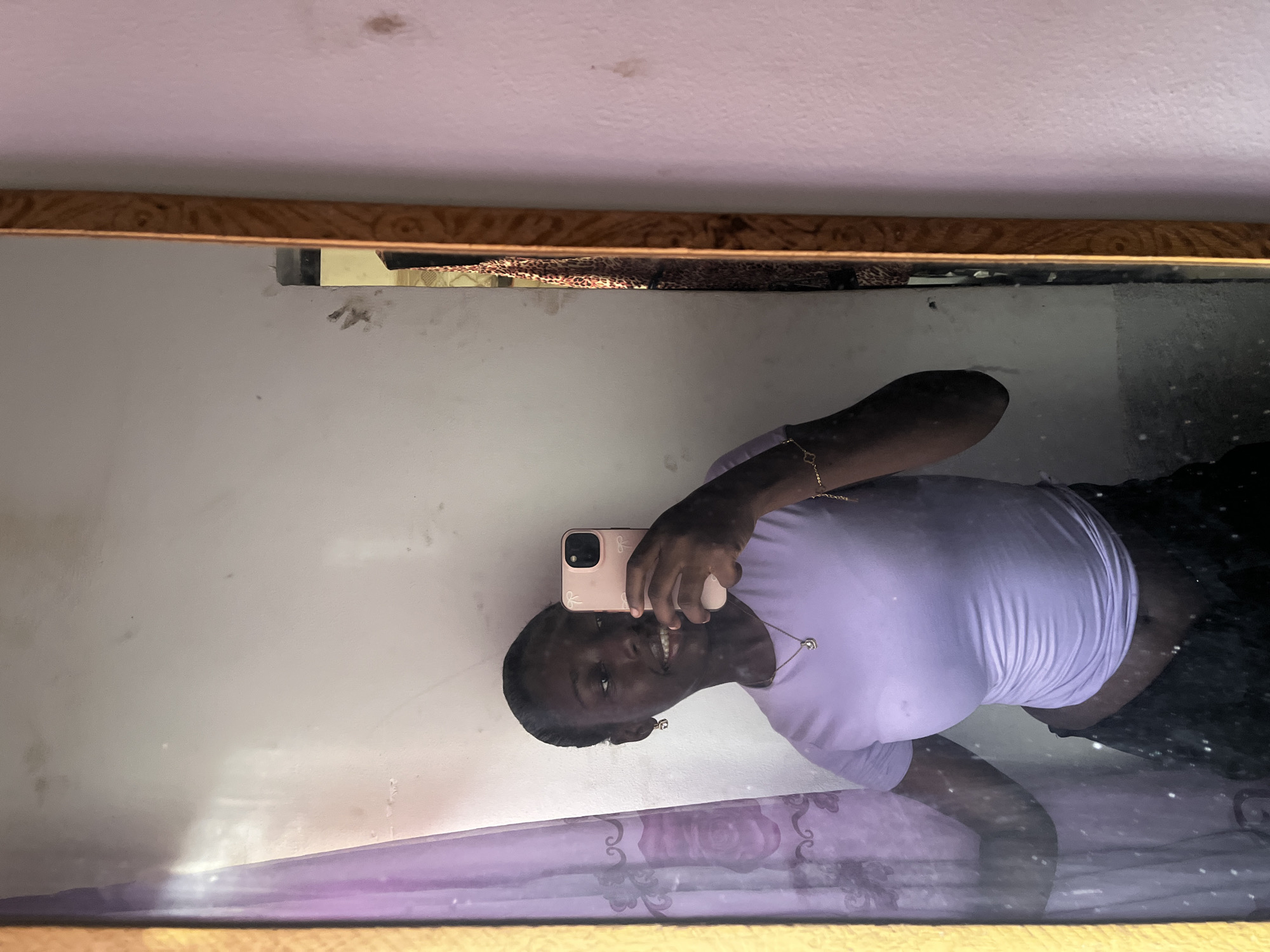 A woman is taking a selfie in a mirror. The woman has dark skin and is wearing a light purple t-shirt. She is holding a pink smartphone. The mirror has a gold-colored frame. The reflection shows a white wall and a patterned purple fabric, possibly a bedsheet. The background suggests an interior setting. The location is unknown, and there is no information provided to suggest the city is Abuja, Nigeria.