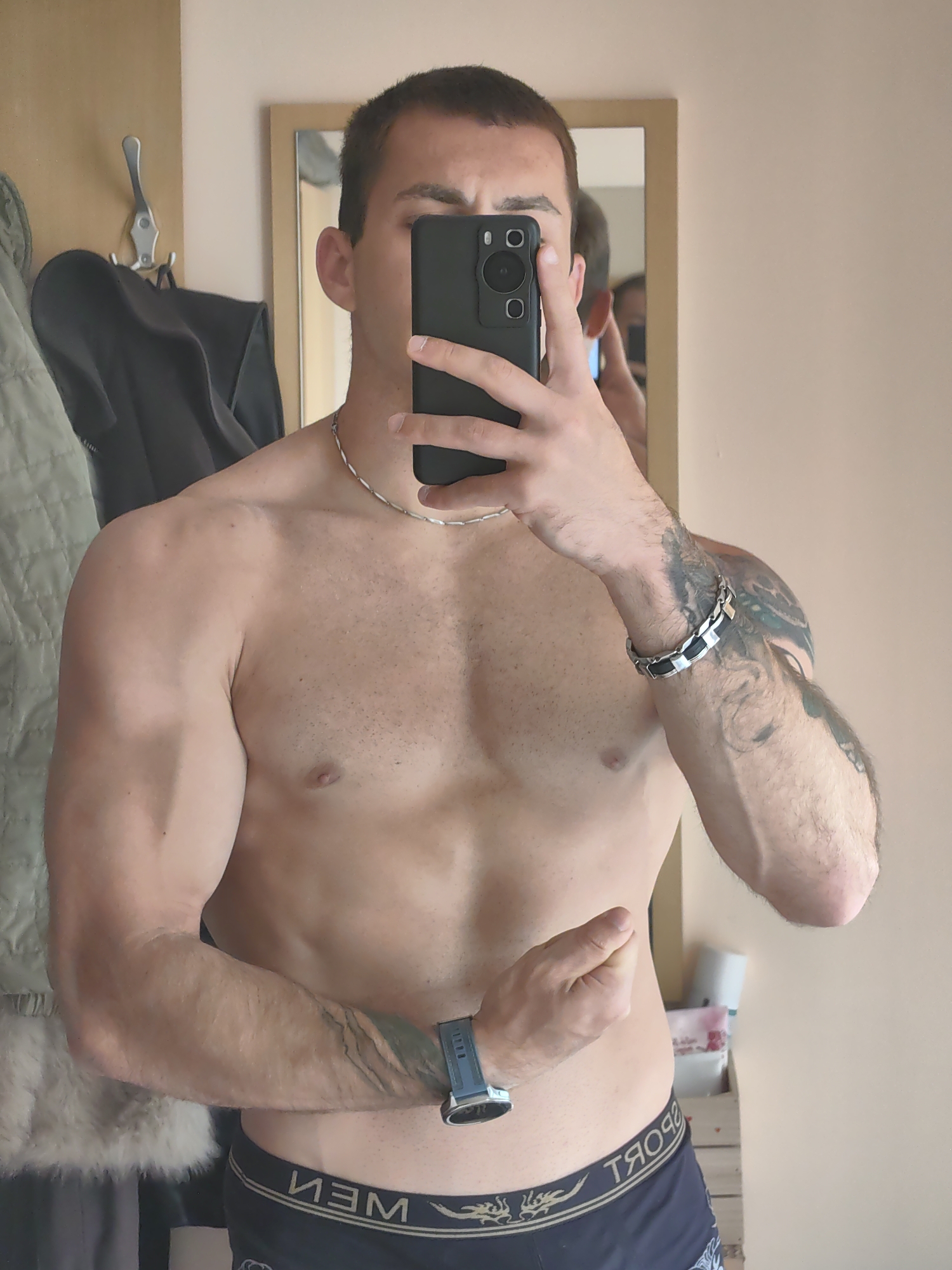 A shirtless man is taking a selfie in a mirror. He is wearing black underwear with "MEN" printed on the waistband. He has a watch on his left wrist and a bracelet on his right wrist. He has tattoos on his right arm and left forearm. In the background, there are clothes hanging on a hook, a mirror, and a shelf with toiletries. The location is not specified in the image.