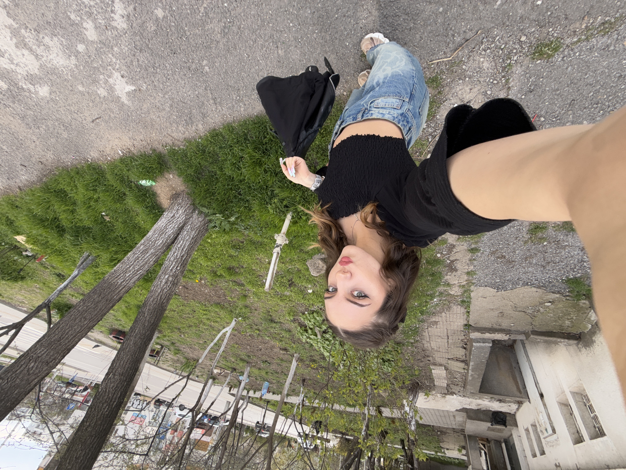 A selfie-style photograph taken outdoors in what appears to be an urban, somewhat dilapidated area, possibly in Sofia, Bulgaria, given the Eastern European aesthetic. The camera is held at an angle, capturing a young woman. She is wearing a black crop top, light blue jeans, and light-colored shoes. A black bag is on the ground near her. The background features cracked asphalt, patches of green grass, trees (some bare, some with budding leaves), and a building with deteriorating concrete. In the distance, there are cars and other signs of city life. The image is rotated, giving a disorienting perspective of the urban environment.