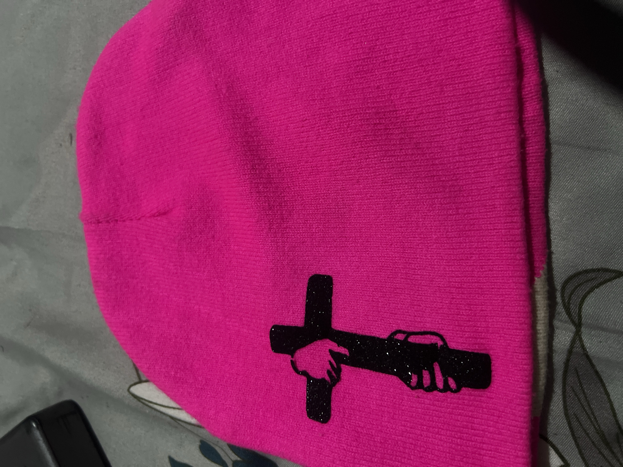 A close-up, overhead shot shows a bright pink beanie lying flat on a grey and white patterned surface. The beanie has a black glittery graphic of a cross with hands holding it, positioned on the left side of the beanie. The graphic is detailed and appears to be an appliqué. The beanie itself is made of a ribbed knit material. The background is out of focus and appears to be a textured fabric, possibly a bedsheet or blanket, with a dark object visible in the upper left corner, which is also out of focus. The lighting is dim and seems to be coming from above and to the right, casting faint shadows. There are no discernible landmarks or features that would suggest a specific location within Jos, Nigeria. The image focuses solely on the beanie and its design.