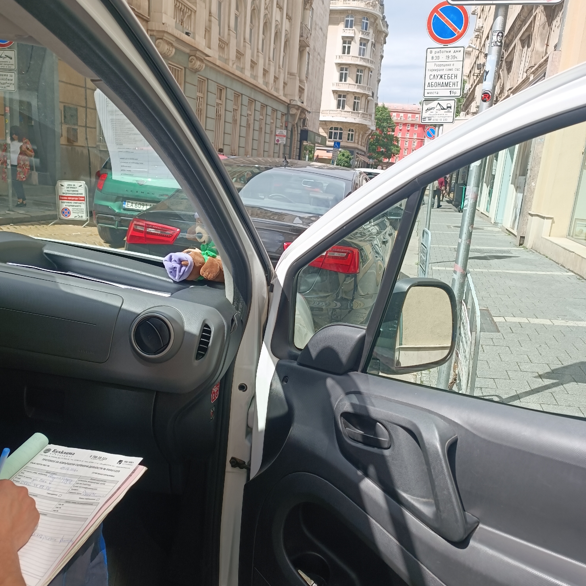 The image captures a first-person view from inside a vehicle, looking out onto a sunny city street in what appears to be Sofia, Bulgaria, given the Bulgarian text on signs and documents. The scene is bright, suggesting daytime with clear weather.
In the foreground, part of the vehicle's dark gray interior is visible, including the dashboard with air vents and the driver's side door panel. A person's hand is holding a clipboard with a document, likely a "ПРОТОКОЛ ЗА ИЗВЪРШЕНИ СЕРВИЗНИ ДЕЙНОСТИ" (Protocol for performed service activities) from "Булклима" (Bulklima). Handwritten notes like "Офис, София" (Office, Sofia) are visible on the document. On the dashboard, two small plush toys, one brown and one green with a purple object, are placed.
Through the open door and windshield, a paved street and sidewalk are visible, lined with multi-story buildings of classical European architecture. Several cars are parked on the street, including a green one and a black Audi (license plate `EA 6091`) whose rear is reflected in the vehicle's interior. A lamppost stands on the sidewalk, adorned with a small sticker of a person's face.
Multiple street signs are prominent. One "No Stopping and Parking" sign (red circle with a diagonal line) specifies conditions: "В работни дни 8:30 - 19:30ч." (On working days 8:30 - 19:30), allowing parking "само със СЛУЖЕБЕН АБОНАМЕНТ" (only with SERVICE SUBSCRIPTION) for "1бр." (1 space). Another sign warns of a "ЗОНА ЗА СИГУРНОСТ С ВИДЕОНАБЛЮДЕНИЕ" (Security Zone with video surveillance) and reiterates the parking ban. A third sign indicates "ВАЖИ ЗА ОМАБИЛИ" (Valid for disabled). Contact numbers for violators (`0700 13 233`) are also listed. In the distance, people can be seen walking on the sidewalk.