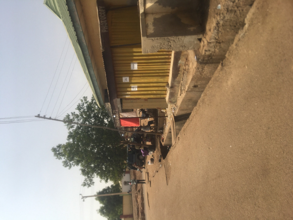 The image captures a street scene in Pindiga, Nigeria, under a clear, bright sky. An asphalt road occupies the left foreground, bordered on the right by a concrete drainage ditch and an unpaved earth shoulder.

Along the right side, a single-story commercial building features a light green corrugated metal roof and a light-colored facade. Its front section is composed of yellow corrugated metal shutters, displaying at least two white rectangular signs. The word "PHOTOCOPY" is stenciled on a brown band above these shutters. Adjacent to this structure on the left, another building also displays "PHOTOCOPY" and has an entrance partially covered by a red fabric.

In the mid-ground, a dark-colored SUV is parked off the road beneath a large green tree. Approximately five individuals are observed gathered around this vehicle. To the left of the SUV, a lone person in light-colored attire stands upright on the shoulder. Near the red fabric and a dark-colored motorcycle, at least two individuals are seated on low stools, with multiple white buckets visible on the ground.

Several utility poles stand throughout the scene, supporting multiple overhead wires that traverse the frame. The background consists of additional trees and distant structures under the hazy sky.