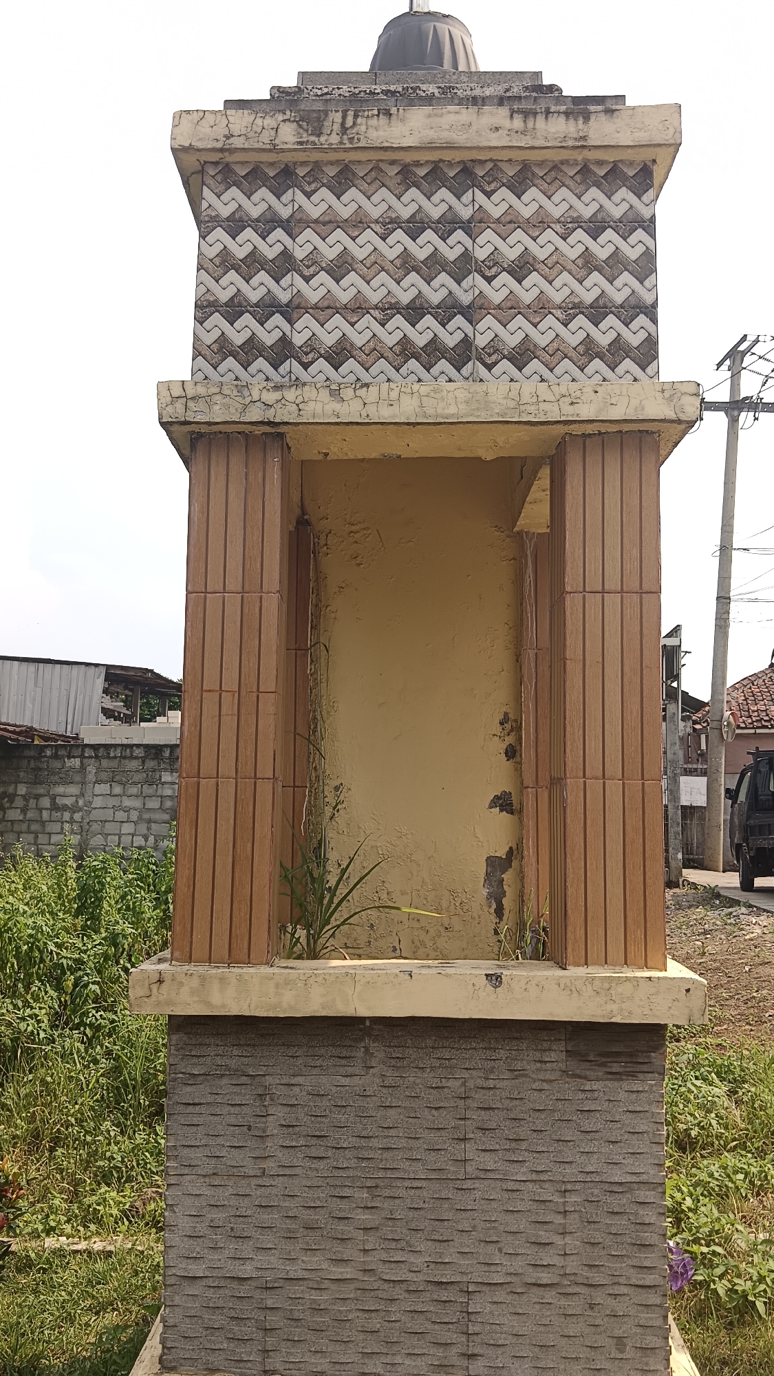 A weathered, decorative structure stands in Kemang, Indonesia, against a bright sky. The structure is rectangular, with a base clad in gray, horizontal stacked stone tiles. Above this is a section with a pale yellow, textured wall set between four fluted wooden pillars. The top of this section is capped by a beige cornice with visible cracks and peeling paint. The upper portion of the structure is decorated with a repeating pattern of diamond shapes created by alternating cream and dark brown tiles. A small, dark dome crowns the entire structure. Green grass and foliage grow around the base of the monument. In the background, to the right, a power line pole and a dark-colored vehicle are visible, along with rooftops of houses. To the left, a low concrete block wall and more greenery are present. The overall scene suggests an outdoor urban or semi-urban environment.