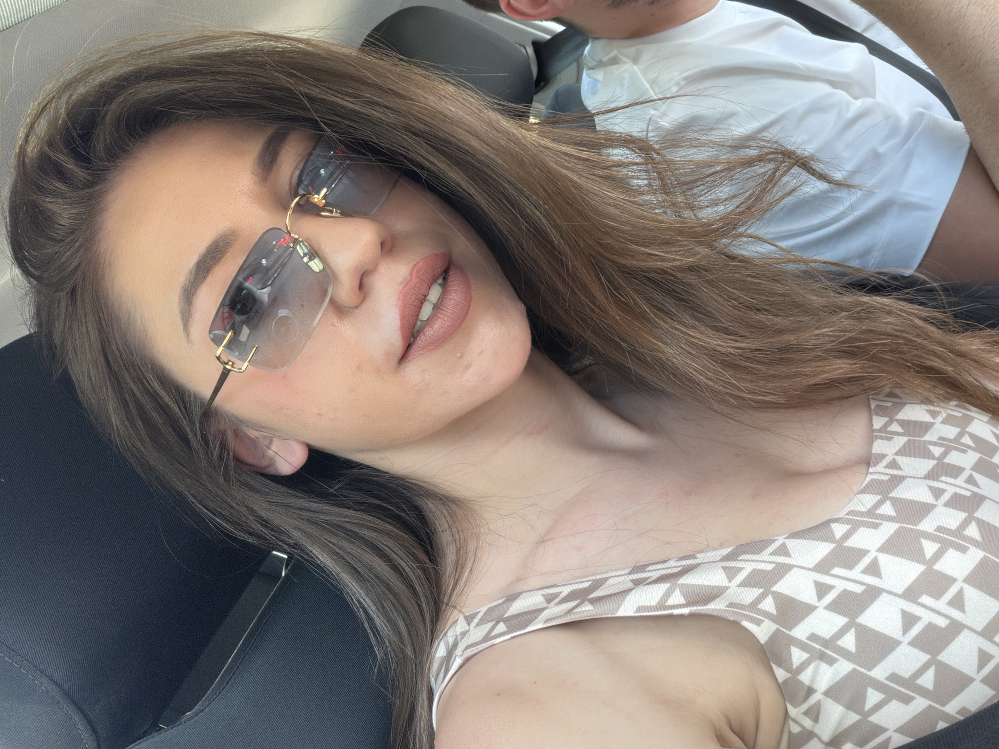 The image shows a close-up, over-the-shoulder selfie of a woman with long, brown hair, wearing sunglasses and a patterned top. She appears to be sitting in a car. The woman is looking at the camera with a slight smile. The background is blurred, but suggests an indoor setting, possibly a vehicle interior. The overall impression is a casual, personal photograph. There are no identifiable landmarks or context that would place this image in Sofia, Bulgaria.