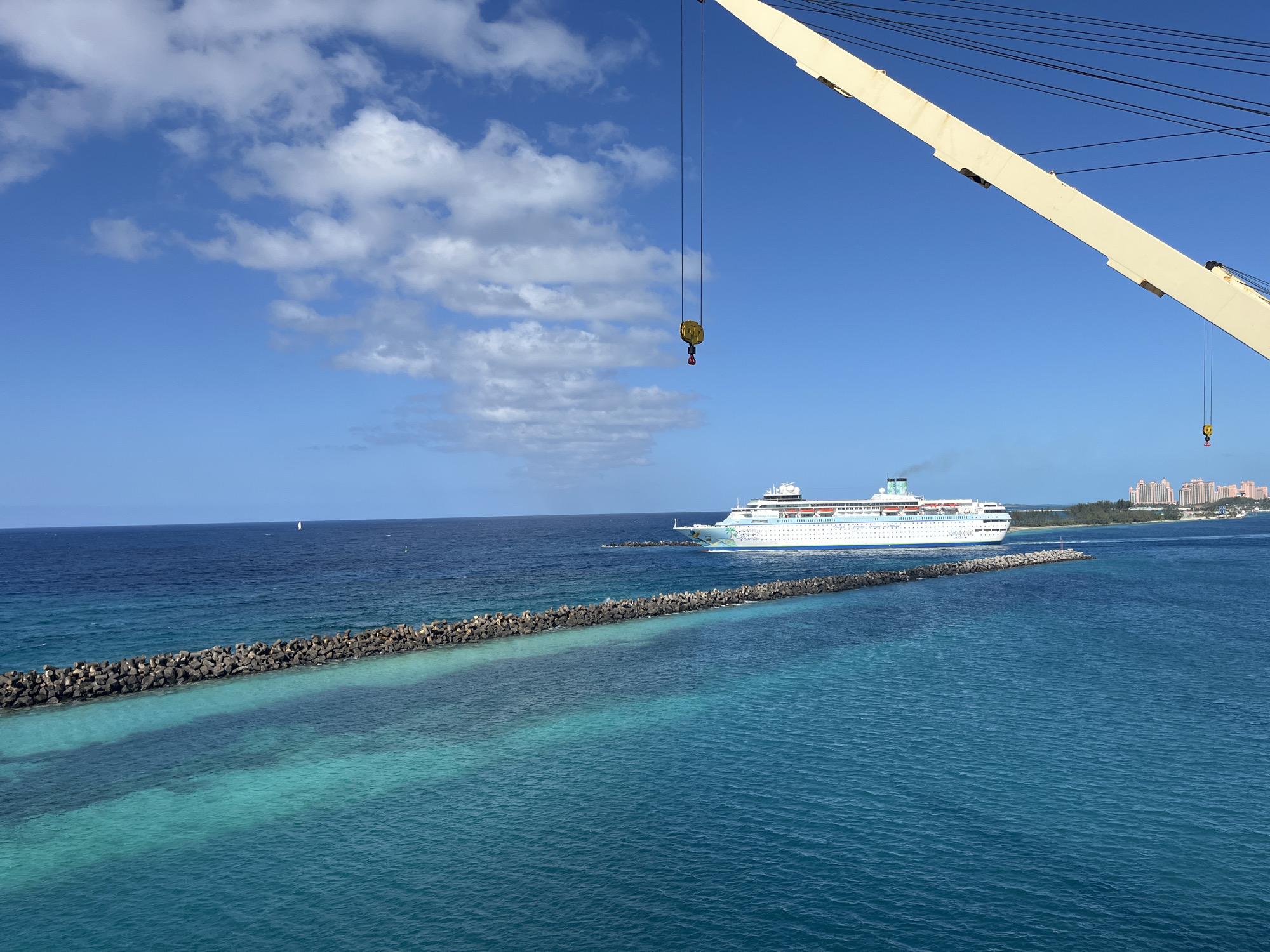 The image shows a wide expanse of turquoise ocean under a partly cloudy blue sky. In the middle distance, a large white cruise ship is sailing past a long, dark-colored rock breakwater. The breakwater divides the ocean into two shades of blue. In the far distance, a line of buildings and trees can be seen along the shoreline. In the foreground, a portion of a white crane is visible. The hook of the crane is suspended in the air. The location is likely coastal, possibly near a port, in an unknown city.