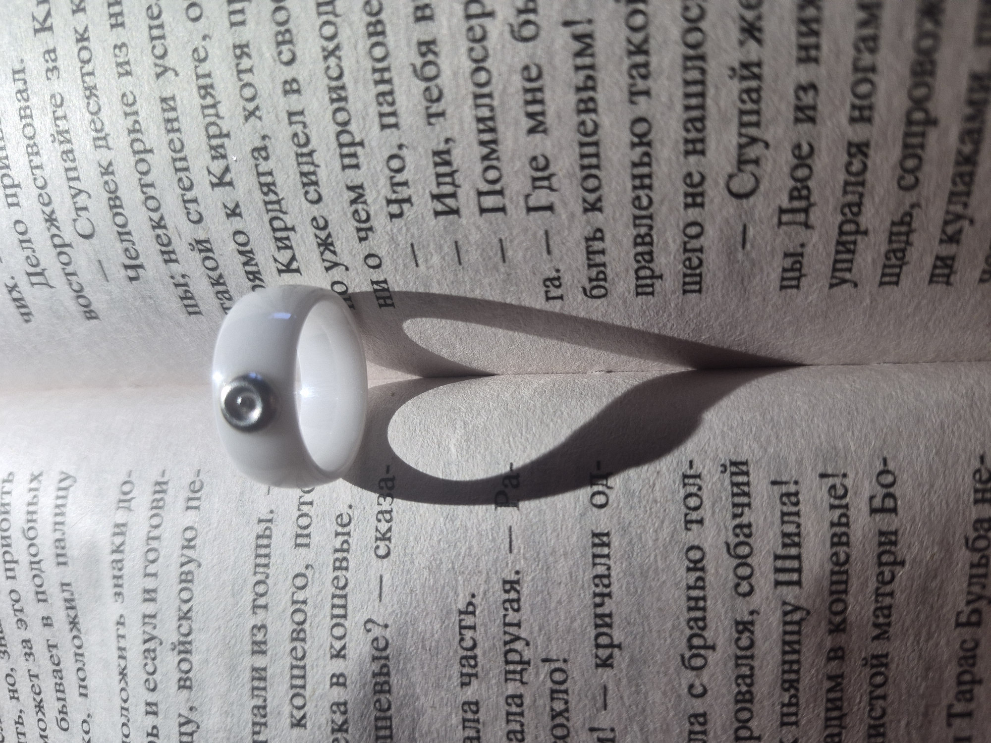 A ring casts a heart-shaped shadow on an open book, its pages filled with Cyrillic text. The ring is white with a metal inset, positioned near the left edge of the frame. The text on the pages is in shades of gray and black. The lighting emphasizes the shadow, creating a focal point.