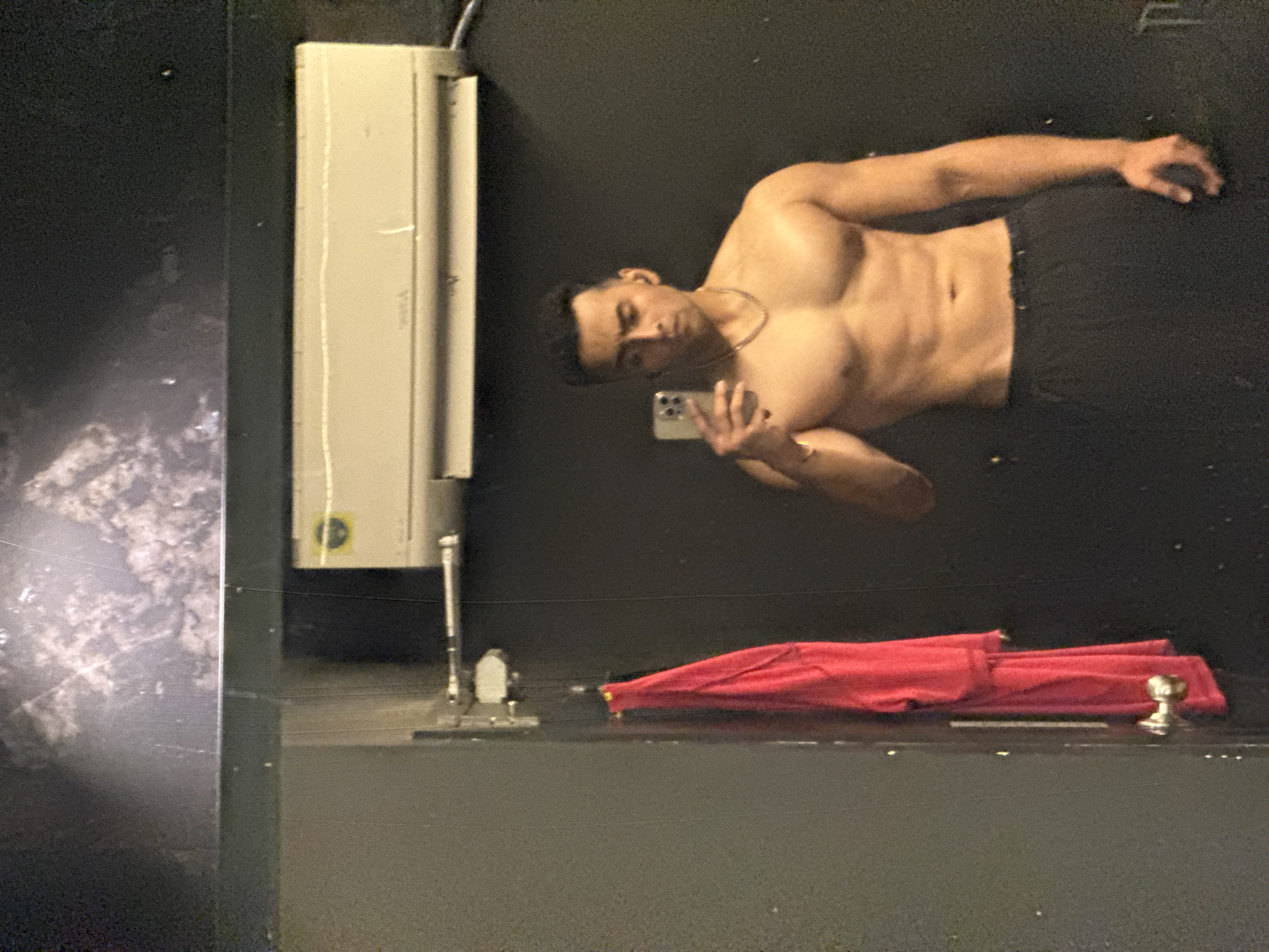 A shirtless man stands in front of a mirror, using a phone to take a picture of himself. A red umbrella lies on the surface in front of the mirror. A light fixture is mounted to the wall on the left. A partial view of a cityscape is visible on the left of the image in the mirror. I can not tell that this is Kolkata.