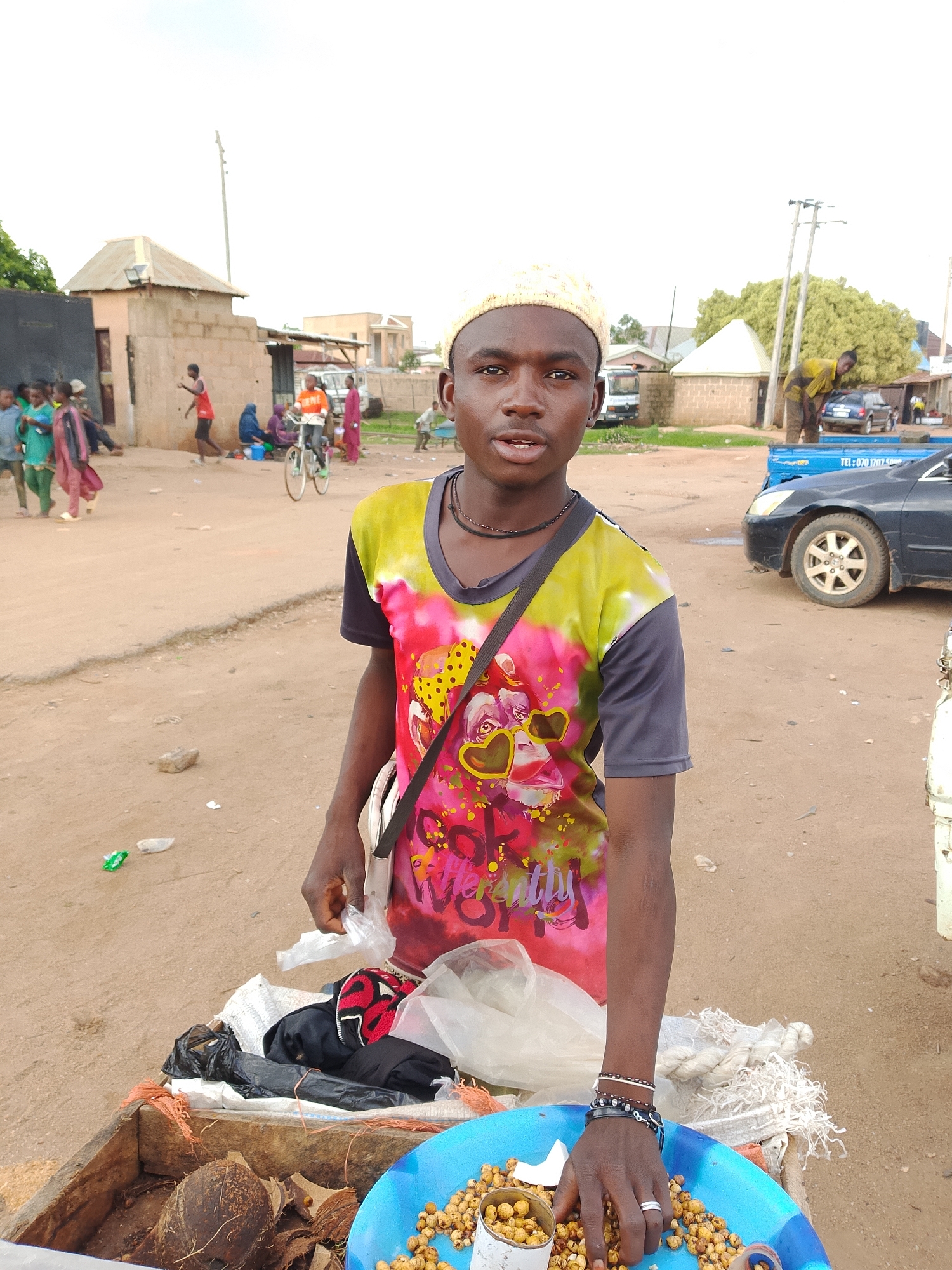 The image shows a young Black male standing behind a cart filled with goods, in an outdoor marketplace in Bukuru, Nigeria. He is wearing a yellow, pink, and black tie-dye t-shirt and a cream-colored cap. A dark crossbody bag is slung over his shoulder. His right hand is reaching into a blue bowl containing small, round, light-colored items, possibly nuts or seeds, and he is wearing several bracelets and a ring on his right hand. The cart contains coconuts and other items covered by black and white plastic bags and fabric. In the background, other people are visible, some walking and others sitting. A black sedan and a blue truck are parked on the dirt road. Buildings with corrugated metal and concrete construction are present in the background. The ground is predominantly dirt, with some scattered debris. The sky is overcast.