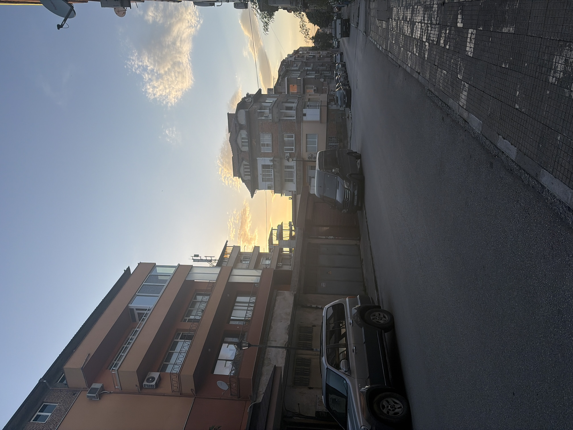 The photograph shows a street scene in Asenovgrad, Bulgaria. The shot is taken from street level, looking down a narrow, paved road lined with multi-story buildings on both sides. The buildings exhibit varying architectural styles and colors, with some displaying weathered facades. Several parked vehicles are visible along the right side of the street, including a light-colored SUV and a black van. Overhead satellite dishes and air conditioning units are mounted on the building exteriors. The sky is visible above the buildings, showing a clear blue with scattered clouds and hints of sunlight suggesting either dawn or dusk. The overall environment appears urban and residential.