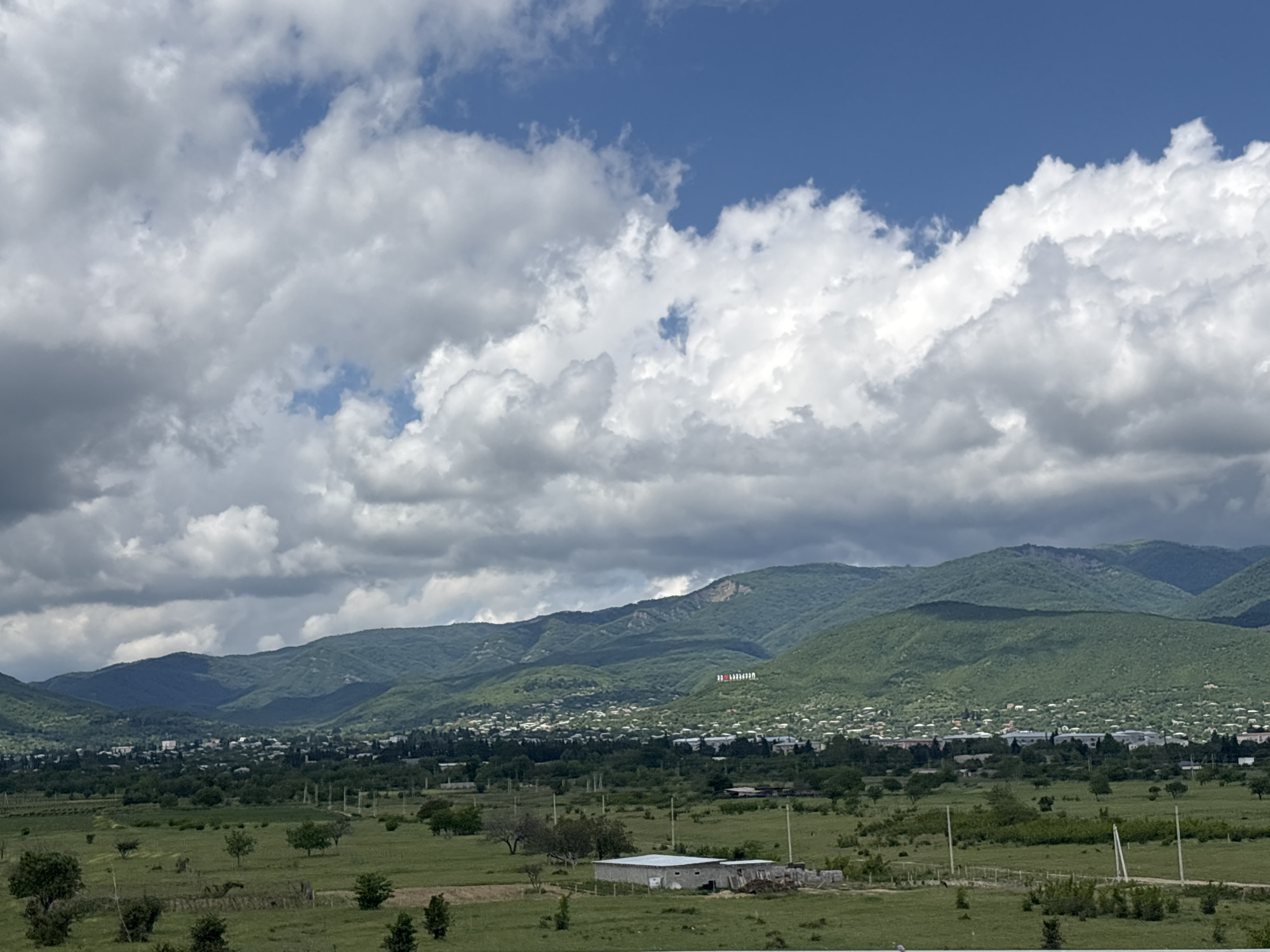 The image depicts a panoramic view of the city of Sagarejo, Georgia, nestled in a valley at the base of lush green mountains. The sky above is a vibrant blue, punctuated by dramatic white cumulus clouds, suggesting fair weather with a possibility of scattered showers.
In the mid-ground, Sagarejo appears as a sprawling settlement with numerous buildings, characterized by a mix of residential houses and some larger structures. The city is spread out, with trees and greenery interspersed throughout. Dominating the hillside behind the city, a large inscription is visible, appearing to be the name of the city in a national flag's colors.
The foreground consists of rolling green fields, dotted with scattered trees and some agricultural land. A simple, light-colored building or structure, possibly a farm building, is situated in the middle of the field. Fencing is visible in sections, and power poles line a dirt road or path that cuts through the landscape. The overall impression is one of a rural or semi-rural setting in a picturesque mountainous region. There are no people visible in the image.