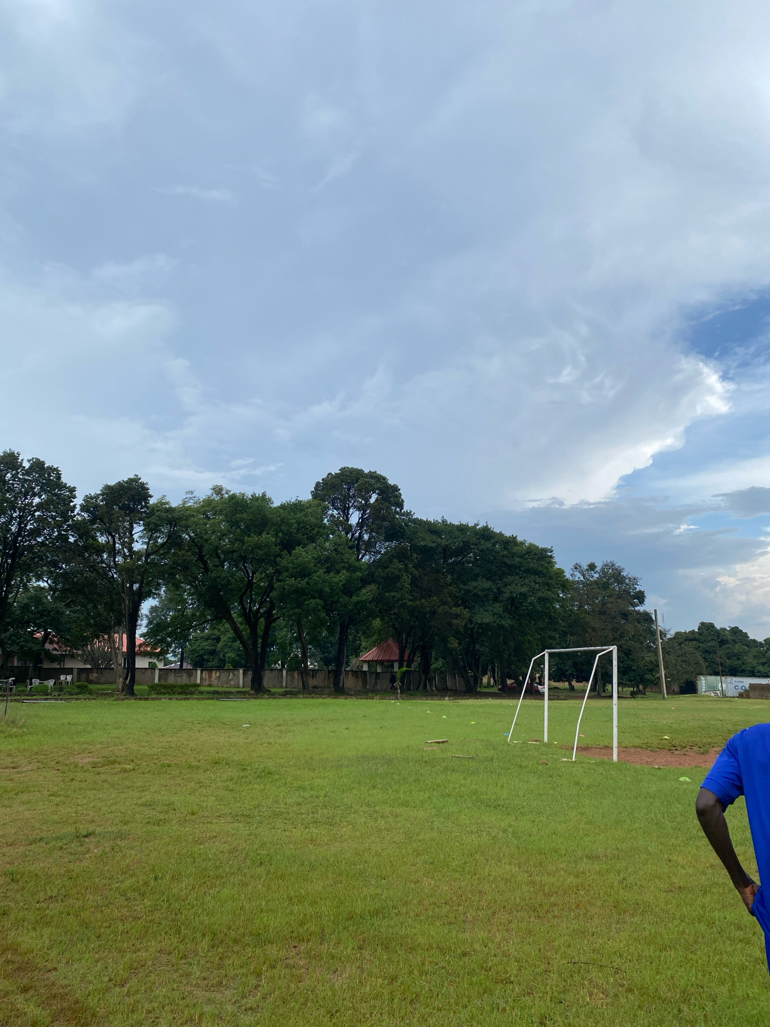 The image depicts an outdoor scene on a grassy field, possibly a sports ground, under a cloudy sky. In the foreground, to the right, the back of a person wearing a blue shirt is visible, with their arm slightly bent and their hand near their hip. The field is covered in green grass, with some sparse patches and irregularities. In the middle ground, a white, rectangular soccer goal is set up. Several small, orange cones are scattered on the grass around the goal. Behind the field, a line of mature, leafy trees forms a backdrop. Beyond the trees, parts of buildings with terracotta-colored roofs are visible, as well as a concrete wall. The sky is filled with a mix of gray and white clouds, with some lighter blue patches suggesting the presence of the sun. The overall impression is one of an overcast day in a semi-urban or park-like setting. While the specific location in Jos, Nigeria cannot be confirmed solely from the visual information, the presence of lush vegetation and the style of the buildings might be consistent with such a locale.