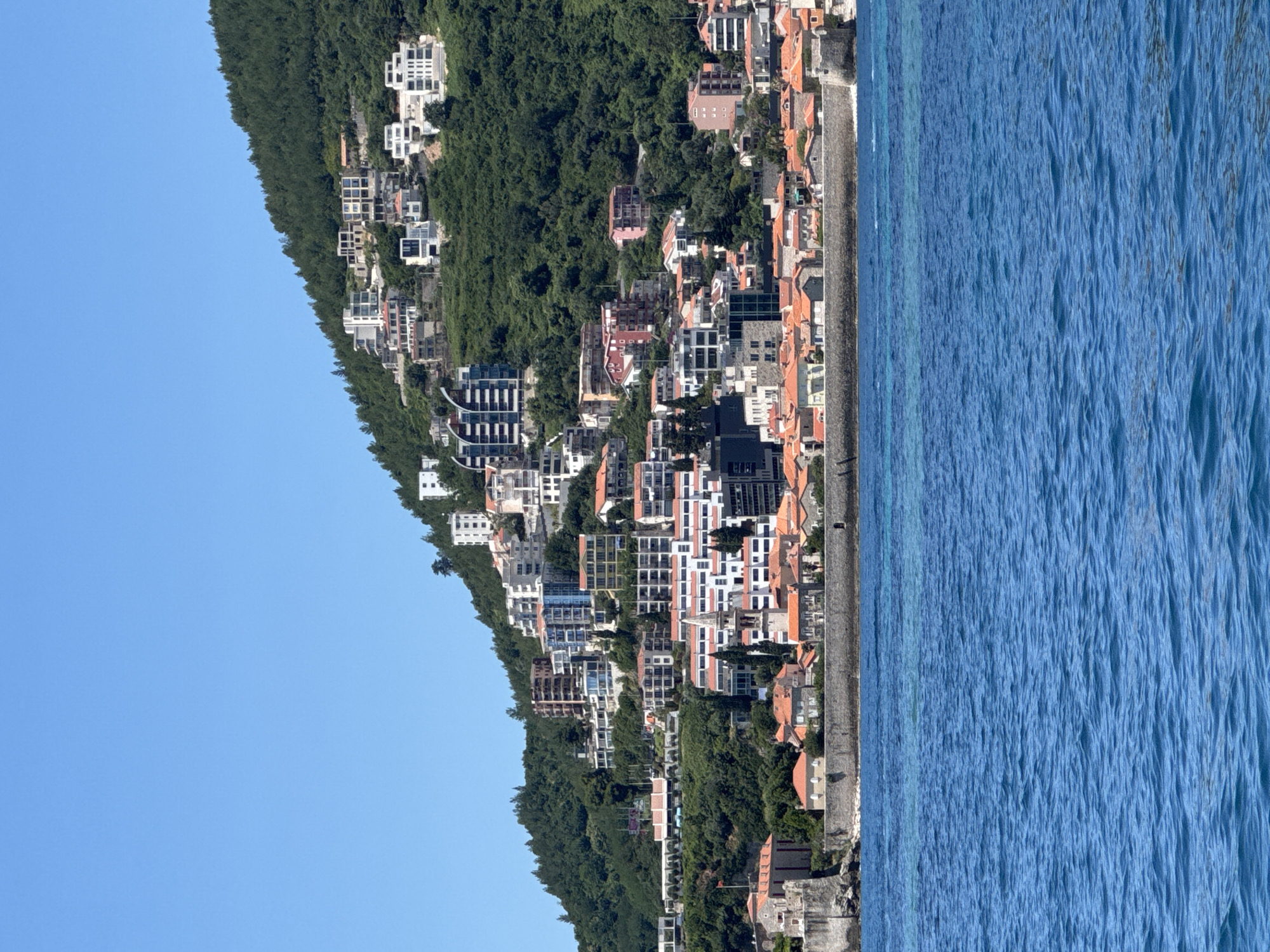 This media file presents an aerial or elevated view of a coastal town, identified as Budva, Montenegro, nestled between a densely forested hillside and the clear blue waters of the Adriatic Sea.
The image, when properly oriented, shows a steep, verdant hill on the left, covered extensively with lush green trees. Buildings are constructed into and along this hillside, descending towards the coastline. The architecture is diverse, featuring numerous multi-story structures. Many buildings are light-colored, predominantly white or off-white, with some showcasing traditional red-tiled roofs. Modern architectural elements, including contemporary designs with glass windows and balconies, are also visible, indicating a mix of historical and newer developments. The town appears densely built, with structures extending significantly up the slope.
To the right, the calm, vibrant blue sea dominates a large portion of the frame. The water displays subtle textural variations and lighter hues closer to the shore, suggesting shallows or a beach area. A distinct coastline separates the urban landscape from the sea, appearing as a narrow strip of land, possibly a promenade or a small beach, lined with buildings.
The scene is captured on a bright, clear day, indicated by the brilliant blue sky, which occupies the upper-left quadrant, and the strong, direct sunlight illuminating the town and water. No specific people or activities are discernible in the image, offering a tranquil, panoramic view of the coastal settlement. There is no visible text within the image.