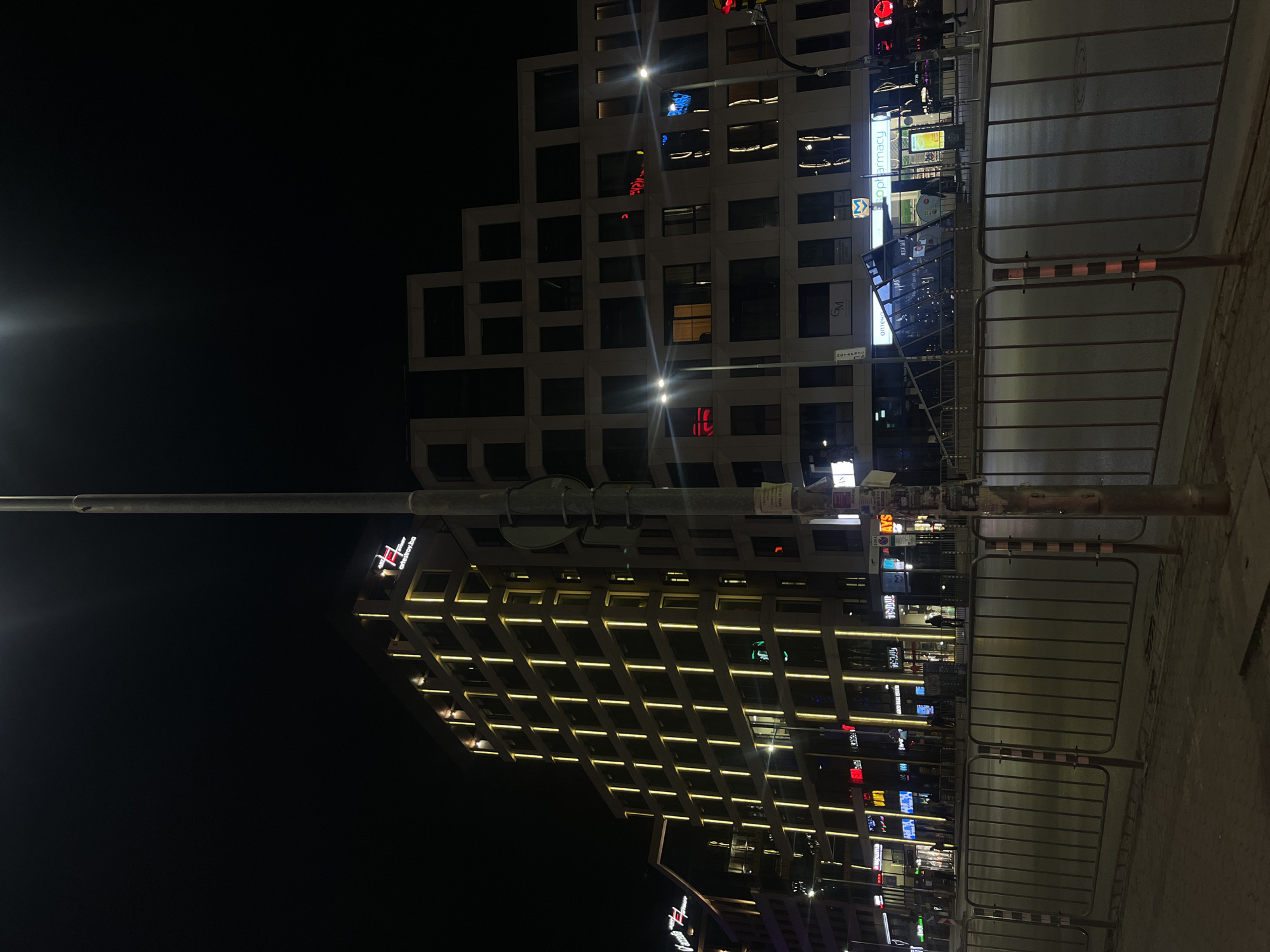 The image depicts a night scene in Sofia, Bulgaria, featuring two modern buildings, a street lamp, metal barriers, and the adjacent sidewalk. The building on the left is illuminated with rows of horizontal lights, and a hotel name "Anel hotel" is prominently displayed. The building on the right has a unique architectural design, with a stepped facade of rectangular windows, some of which are illuminated. The sky is dark, and the street is dimly lit. The metal barriers line the sidewalk. There are no people clearly visible in the image.