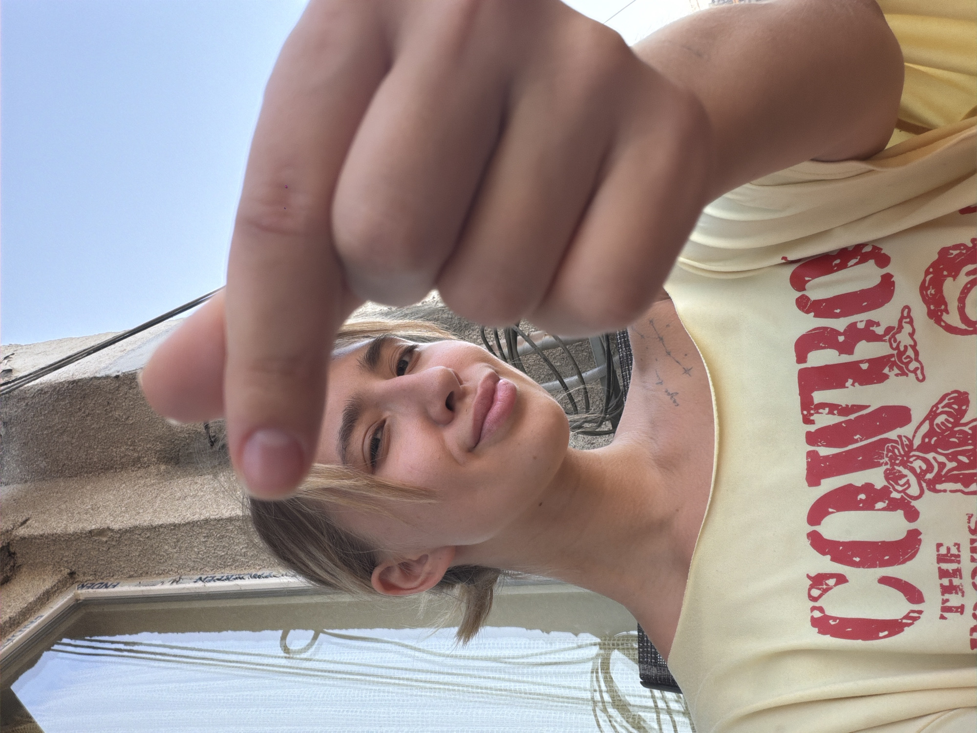 A young woman with blonde hair pulled back into a ponytail and bangs is shown in a close-up selfie. She is wearing a light yellow t-shirt with a red graphic that reads "COWBOY THE BOOTS". Her right hand is held up in the foreground, with her thumb and index finger touching in a gesture. She is smiling and looking at the camera. The background shows a building with a window and a clear blue sky. The image appears to have been taken outdoors, possibly on a balcony or patio. The woman has a tattoo on her left shoulder blade. The location of the image is the city of Tbilisi, Georgia.