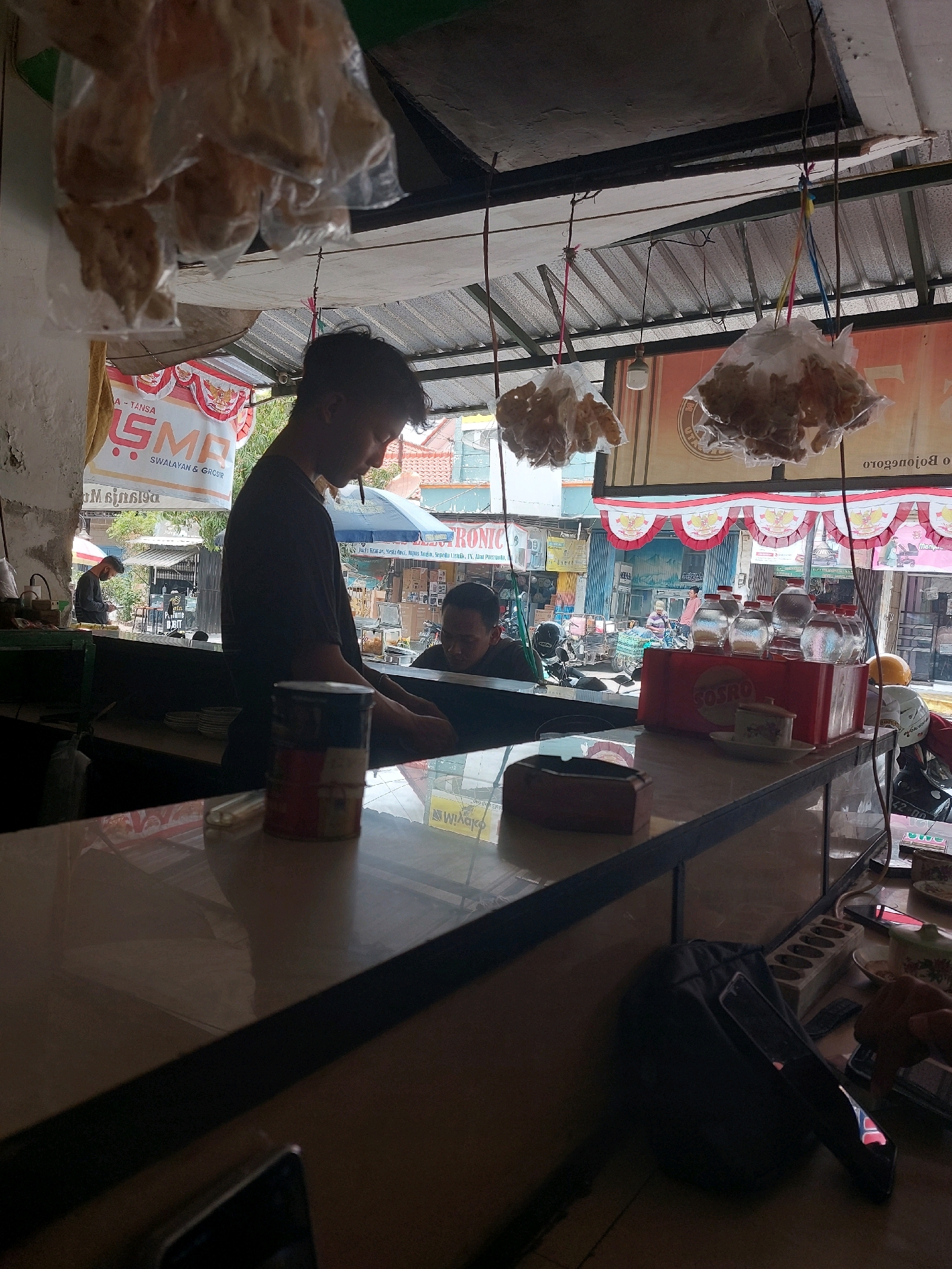 In Sumuragung, Indonesia, an indoor scene depicts a roadside stall with a canopy. Two men are present; one, with dark, short hair, stands behind a counter, appearing to be engaged in his work. The other man is partially visible, seated and looking downwards. The counter area is cluttered with various items including a tin can, a plastic container with bottles, and an ashtray. Hanging from the ceiling are plastic bags filled with food items. The stall's background reveals a street scene with buildings, parked motorcycles, and other stalls, indicating a bustling commercial area. The overall lighting suggests it is daytime.