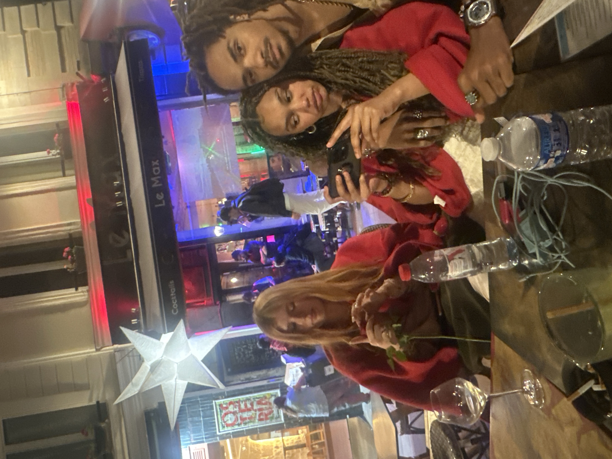This media file captures an evening scene, likely on a street or terrace in Paris, France, outside an establishment named "Le Max." The image appears to be rotated 90 degrees clockwise.
In the foreground, a man and a woman are seated closely together. The man, with dreadlocks and a beard, looks towards the camera with a subtle smile, his head resting on the woman's shoulder. The woman, also with dreadlocks, holds a smartphone in her hands, seemingly focused on its screen, perhaps taking a selfie or viewing content. Both are wearing red tops or jackets. The man wears a watch, and the woman has several rings on her fingers.
To their right, another woman with long blonde hair is seated at a table, also wearing a red top. She is looking down, gently holding a single red rose. On the table in front of her are a plastic water bottle, partially labeled "Aurelia," a clear stemmed wine glass, and another glass containing a dark beverage. Some cables are also visible on the table.
In the background, the entrance and interior of a bar or restaurant are visible. A vertical sign clearly reads "Le Max" and "Cocktails." The interior is brightly lit with colored lights, predominantly red and blue, and several other people are discernible inside, socializing. A large, white, star-shaped paper lantern hangs in the mid-ground. Further back, a framed artwork or sign with some red text, possibly "GALERIE," is seen.
The scene conveys a relaxed, social atmosphere during the evening or night, with people enjoying drinks and company.