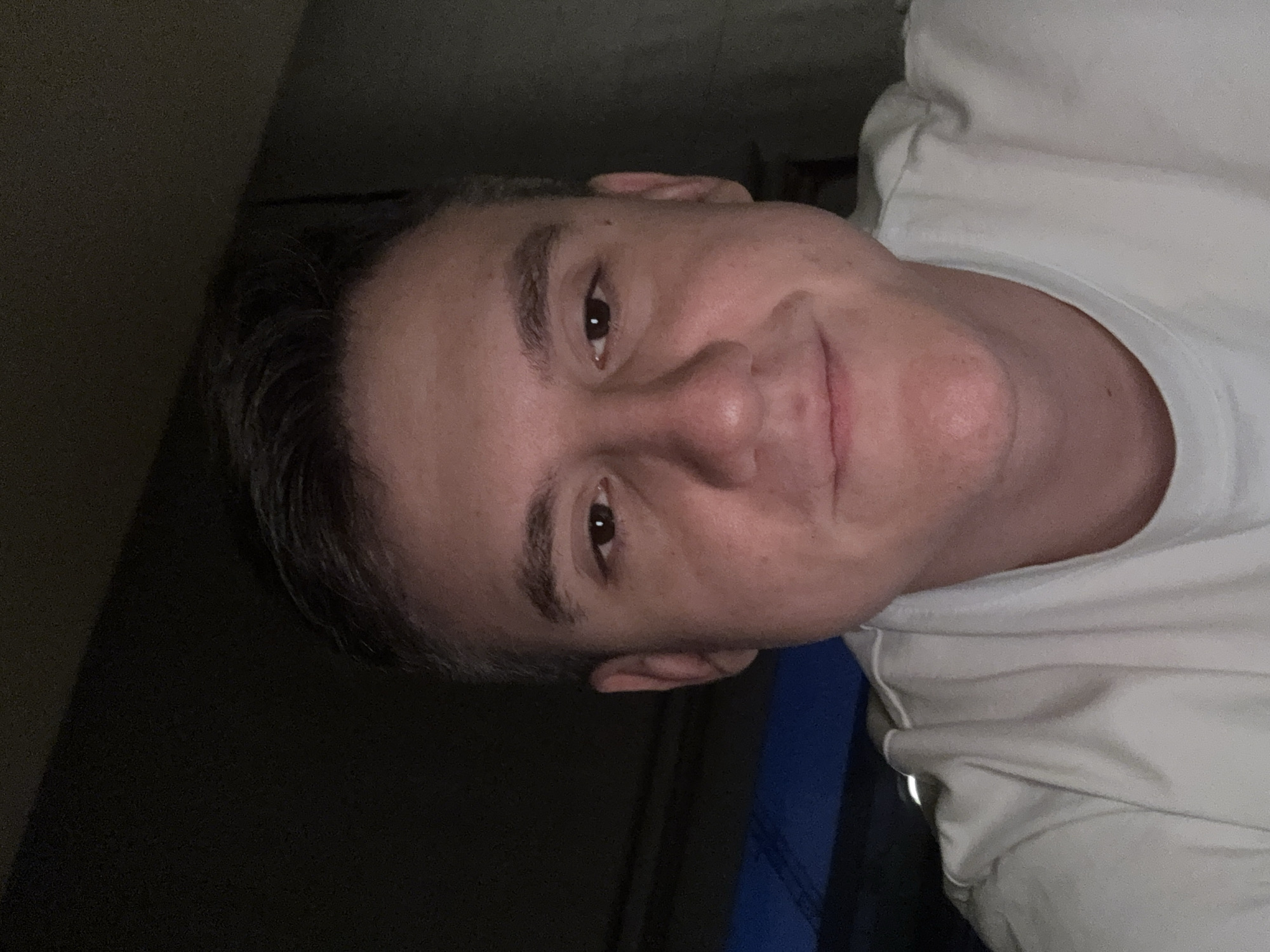 A close-up, slightly tilted shot features a fair-skinned man with short, dark hair. He is wearing a light-colored t-shirt. The background is dark and indistinct, suggesting a poorly lit indoor setting. There is a blue-colored object behind the man's shoulder. The location cannot be determined from this image. There is no information to suggest the location is Warsaw, Poland.