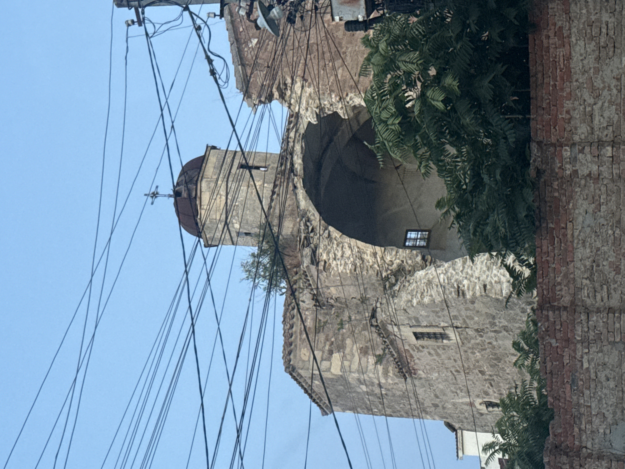 The image shows a damaged stone building, likely a church, in Feodosia, Ukraine. The building has a partially collapsed arched opening and a dome topped with a cross. The exterior walls are made of rough-hewn stone. Overgrown vegetation is visible around the structure. Numerous electrical wires are strung across the sky, obscuring parts of the building and the sky. In the foreground, a low brick wall runs horizontally across the image. The sky is clear and blue, suggesting daytime.