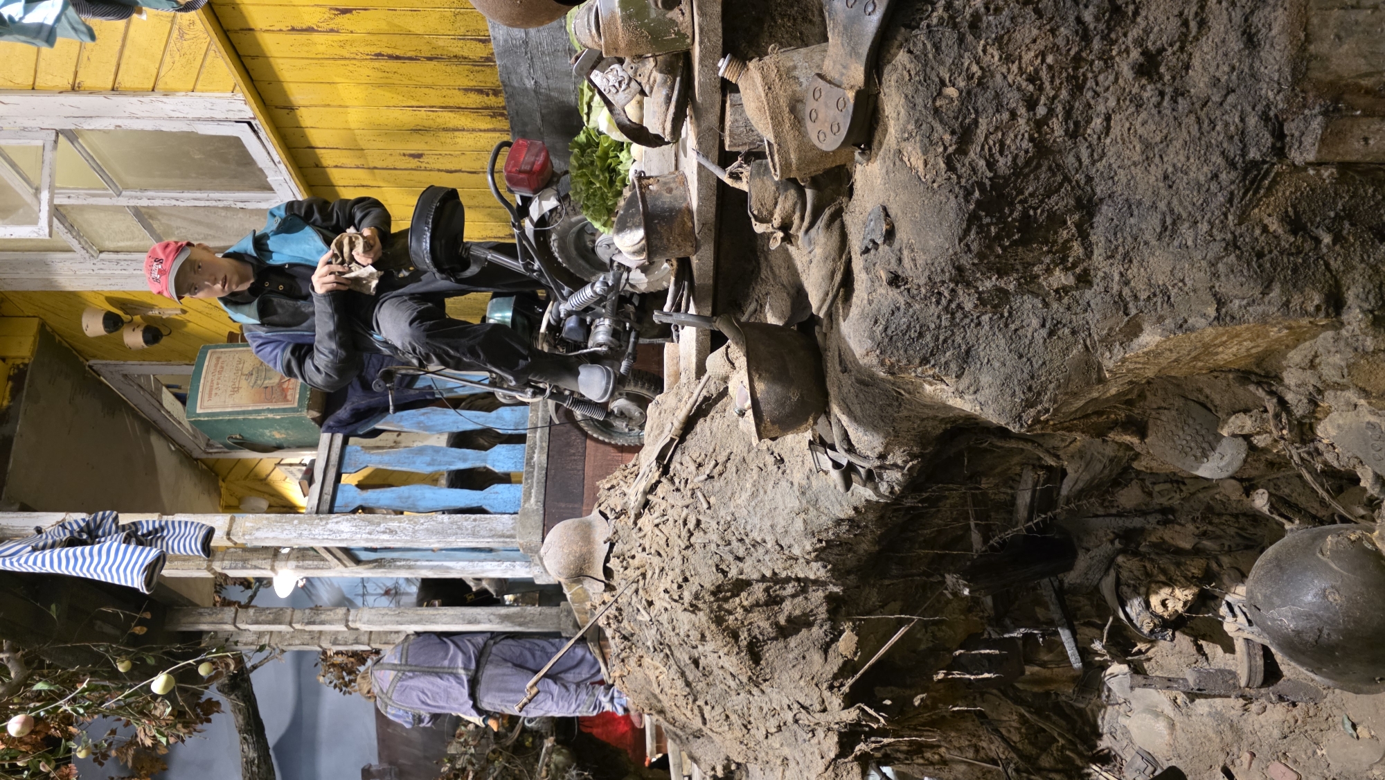 The image depicts a scene that appears to be a museum display or installation. A person wearing a red cap, blue jacket, and black pants is sitting on a blue wooden structure, possibly cleaning or working on a motorcycle part. The setting seems to be a diorama of some kind, with the yellow building in the background resembling a simple structure, possibly a house or shed. To the right and below, there is an area designed to simulate a muddy or earthen environment with various objects embedded in it, including what looks like old military helmets and pieces of equipment. A mannequin dressed in a blue outfit is visible towards the bottom left. The overall impression is that the location may be a history museum or exhibit dedicated to historical events, perhaps military history, located in an unknown city and region.