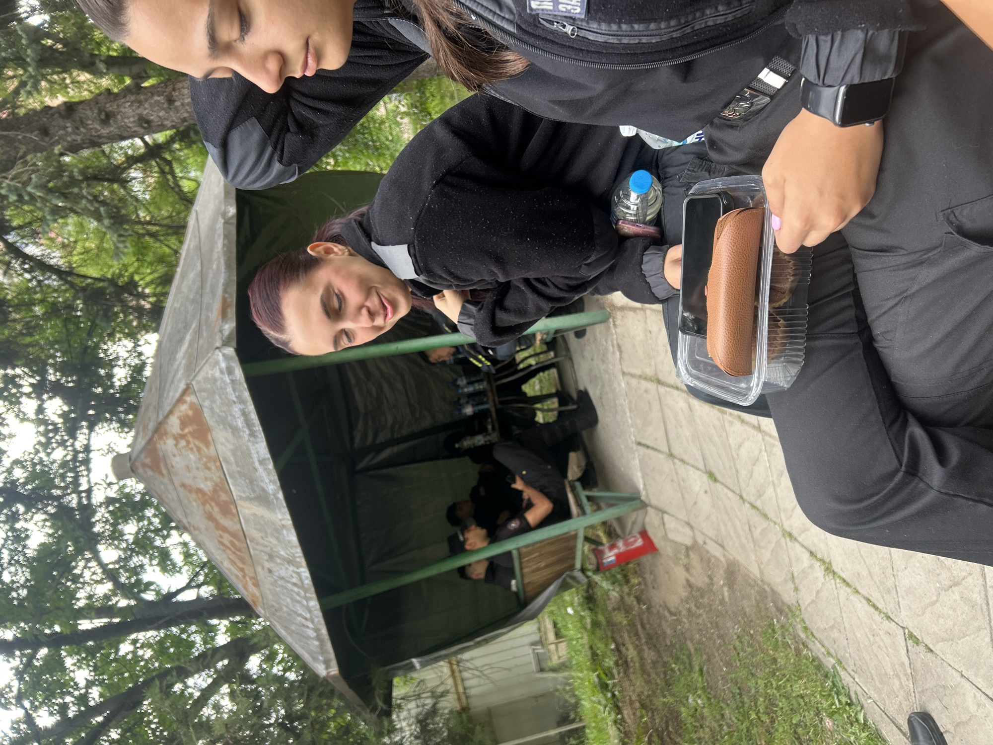 The image shows two women in Sofia, Bulgaria, standing outdoors. One woman looks at the camera and smiles. The other appears to have her eyes closed. Both are wearing black tops and black pants. One of the women holds a clear plastic container with a brown case inside. A water bottle is visible nearby. In the background, inside of a metallic shed, there are other individuals wearing what appears to be uniforms. The shed is outdoors, and the scene appears to be located near a wooded area. The ground is paved with a patterned stone pathway.
