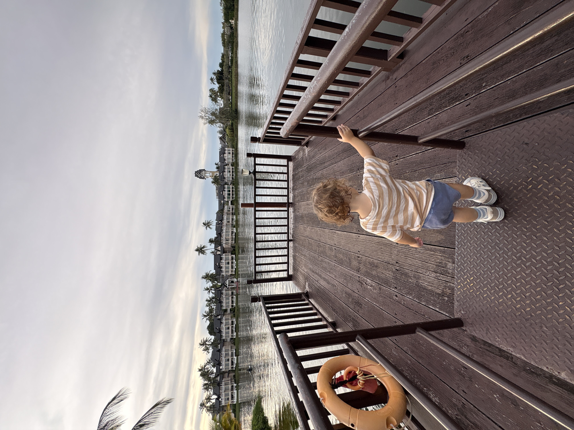 A young child stands on a wooden pier overlooking a body of water. The child is facing away from the camera, wearing a striped t-shirt and blue shorts. The pier is made of dark wood planks and has brown railings on either side. An orange life preserver is attached to the railing on the left. In the background, across the water, are several buildings with dark roofs, resembling a resort or residential complex, with palm trees in front of them. A tall, square tower with a clock is visible in the distance to the right of the buildings. The sky is overcast with pale clouds. The location is likely a waterfront area in Choeng Thale, Thailand, suggested by the architecture and palm trees.