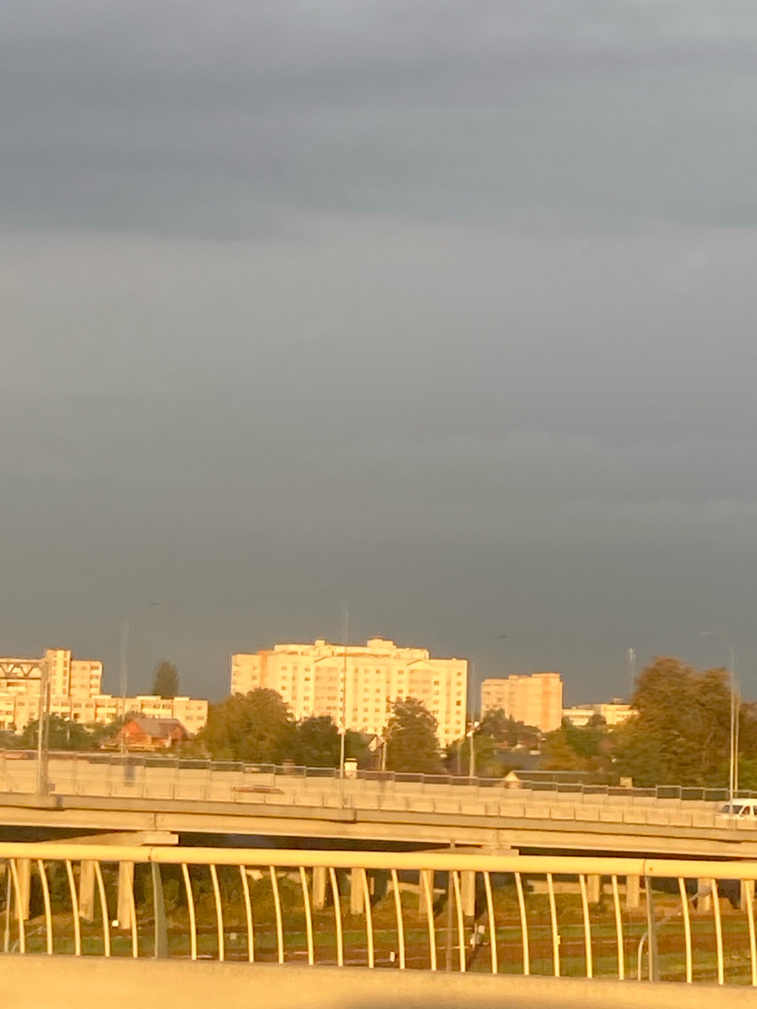 The image captures a view of apartment buildings in the city of Brest, Belarus, under a cloudy sky. In the foreground, a multi-level highway overpass is visible, with a white barrier railing made of vertical bars. The overpass appears to be made of concrete. Beyond the overpass, several tall apartment buildings, primarily light-colored, dominate the middle ground. Some trees and a small red building are also present among the apartment structures. The sky is overcast with a gradient of gray, suggesting a potentially stormy or evening atmosphere, with some hints of sunlight illuminating the buildings from the side. The overall impression is an urban landscape with infrastructure and residential areas.