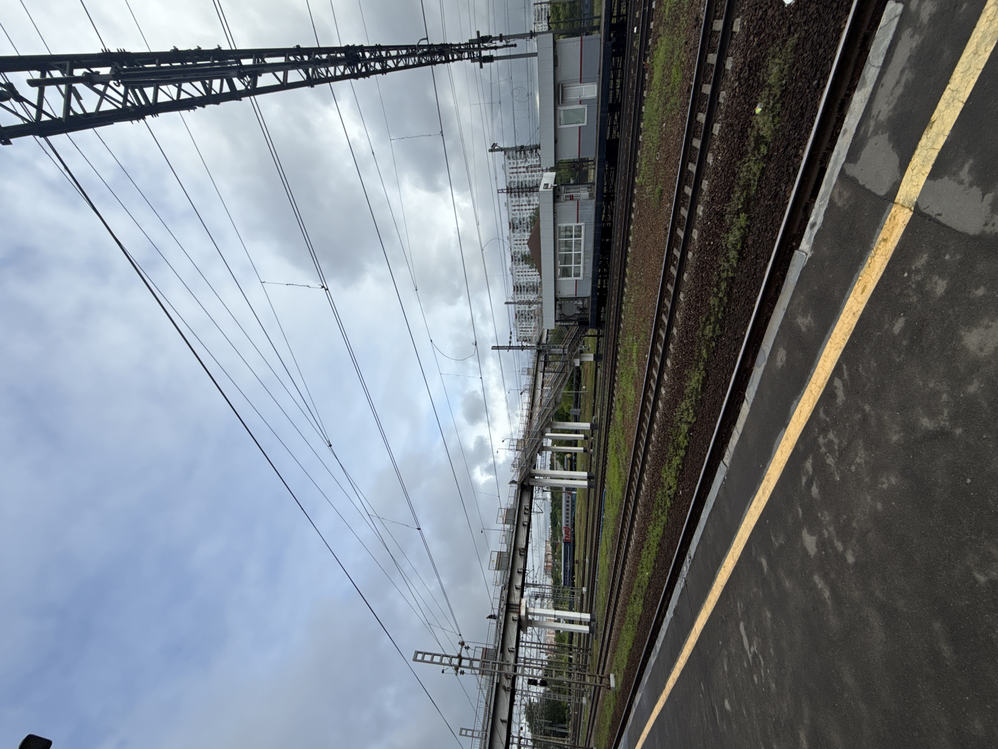 The image shows a railway platform in Moscow, Russia, under an overcast sky. The platform is marked with a yellow safety line and appears wet. Adjacent to the platform are railway tracks with ballast and sparse vegetation. Overhead infrastructure includes electrical lines supported by metal towers. In the background, a pedestrian overpass spans the tracks, and a building can be seen further off, along with high-rise apartments in the distance. No people are visible in the immediate scene.