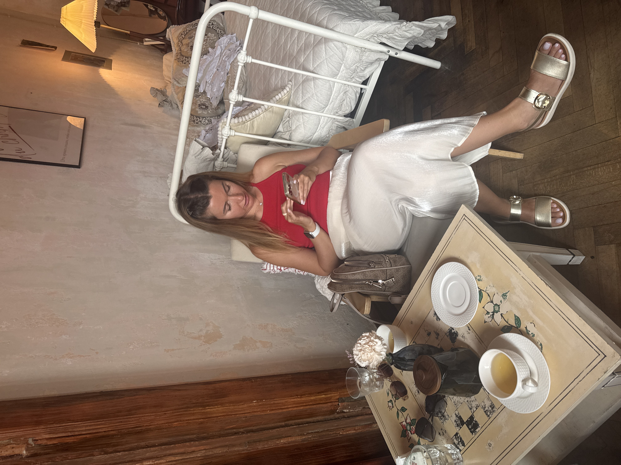 A woman is seated in a chair, looking at her smartphone. She is wearing a red tank top and a flowing, light-colored skirt, with gold sandals. Beside her, a small table holds a tea set, a vase with a flower, and sunglasses. Behind her, a bed with ornate white bedding is visible. The room appears to be in an apartment in Saint Petersburg, Russia. The walls are textured and light-colored, with some peeling paint, and the flooring is dark wood. A lamp with a yellow pleated shade casts a warm glow. A framed poster hangs on the wall.