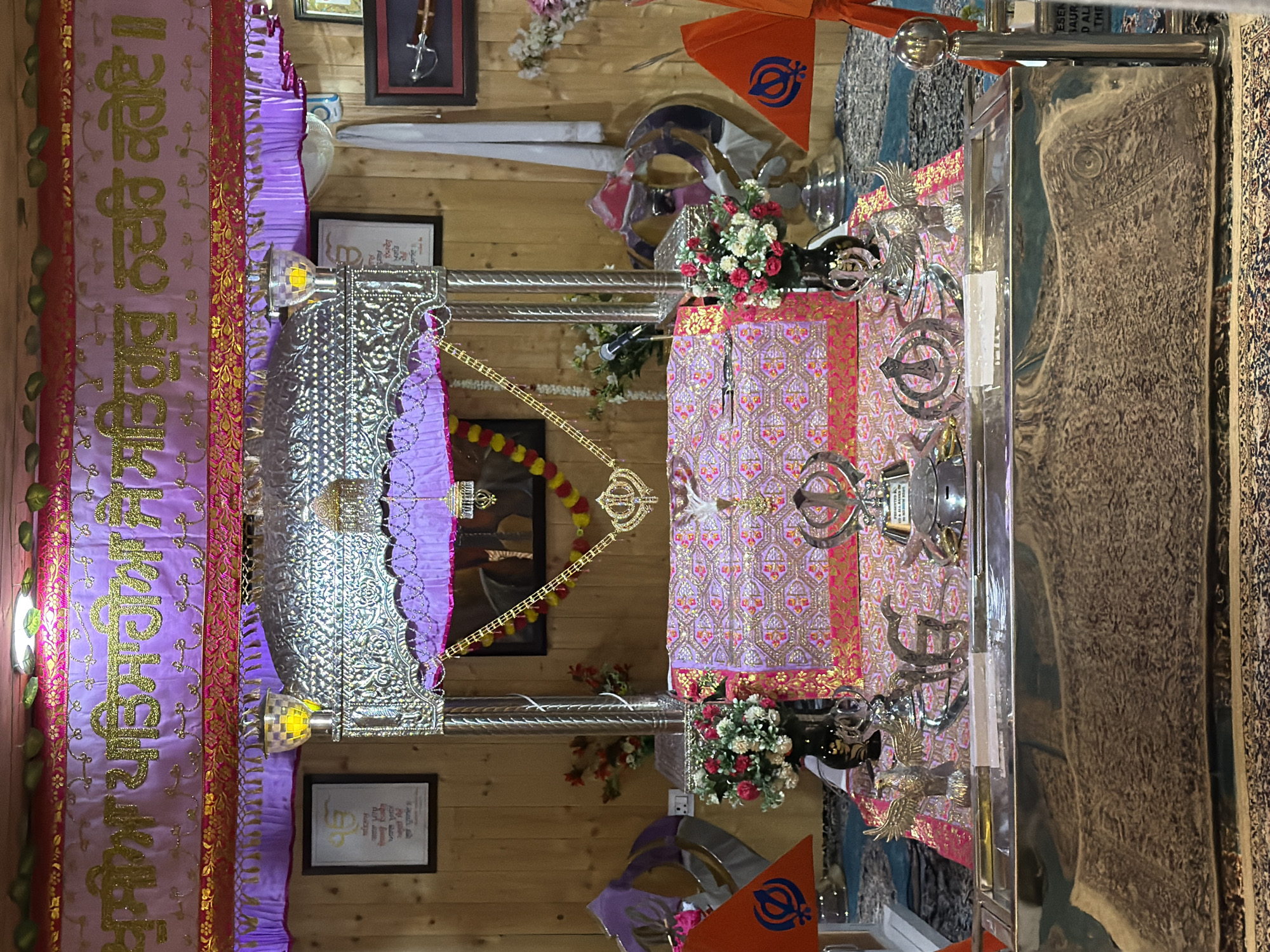 This image shows the interior of a Sikh Gurdwara, likely located in Chumkar, India. The scene is dominated by a raised platform covered in ornate pink and gold fabric, on which rests a large, silver-colored structure resembling a canopy. Beneath the canopy, a book, likely the Guru Granth Sahib, is covered with a patterned cloth.
Various religious symbols are visible, including the Khanda, a Sikh emblem, depicted in silver on the platform and as part of a flag. Two orange flags with Khanda symbols are placed on either side of the platform. Decorative elements include floral arrangements in vases, silver eagles, and metallic finials.
The background consists of wooden paneled walls, adorned with framed pictures and religious texts. A purple and gold banner with Sikh inscriptions hangs across the top of the scene. The lighting appears to be from ceiling-mounted fixtures. There are no people visible in the image, and the scene is static, suggesting a devotional space prepared for religious observance.