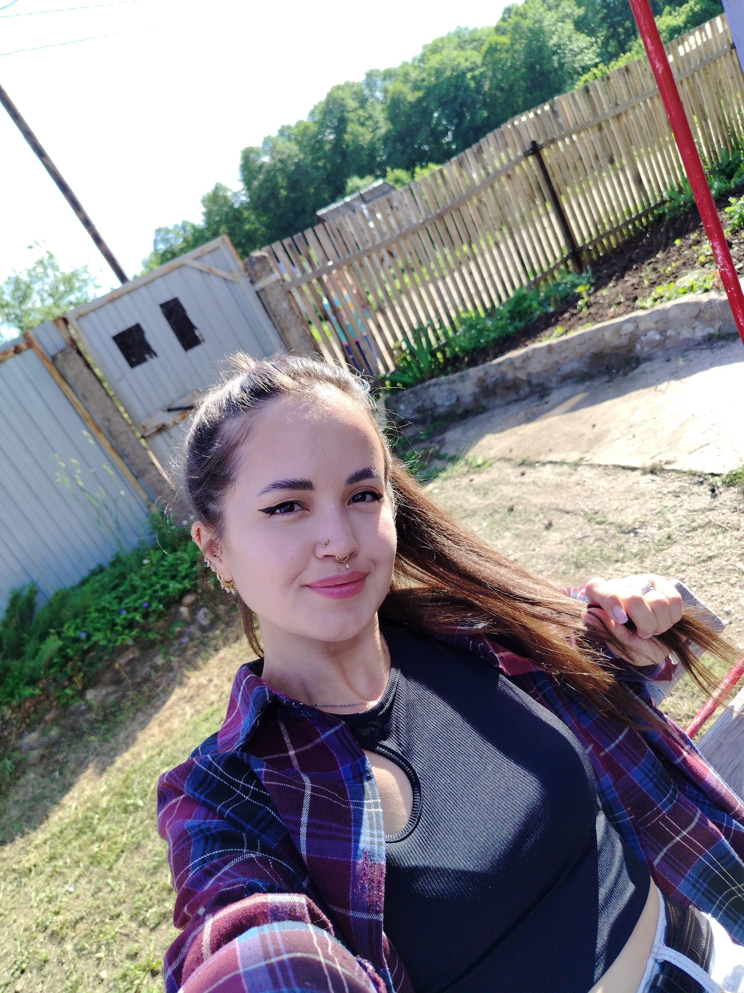 This image, situated in the city of Noviy, Russia, depicts a young woman in an outdoor setting under bright daylight. She is facing the camera with a soft smile, her head tilted slightly. She possesses long, dark brown hair, which is partially pulled back, with loose strands cascading over her left shoulder. Her right hand is visible, lightly grasping a section of her hair. She is wearing a black ribbed top featuring a circular cutout on the chest, layered under an open plaid flannel shirt in shades of purple, blue, and red. A thin silver chain necklace is visible around her neck. Her facial features include dark, well-defined eyebrows, winged eyeliner, and light-colored lipstick. She has multiple piercings: a gold-toned circular septum ring, a small stud in her right nostril, and various studs and hoops with small decorative elements in both ears, including helix, tragus, and lobe piercings. A ring with a prominent clear stone is worn on her left ring finger.
The background consists of a mixed environment. To the left, a light-colored corrugated metal fence or wall, possibly a shed, displays two dark, rectangular openings. To the right, a wooden picket fence runs across the mid-ground. Beyond the fences, dense green trees indicate a vegetated area. The ground foreground is a mix of dirt, sparse grass, and some concrete or paved sections. A red vertical pole or structure is partially visible on the far right, and a utility pole with wires stands in the upper left background. Strong sunlight casts shadows, particularly on the woman's right side and her clothing.